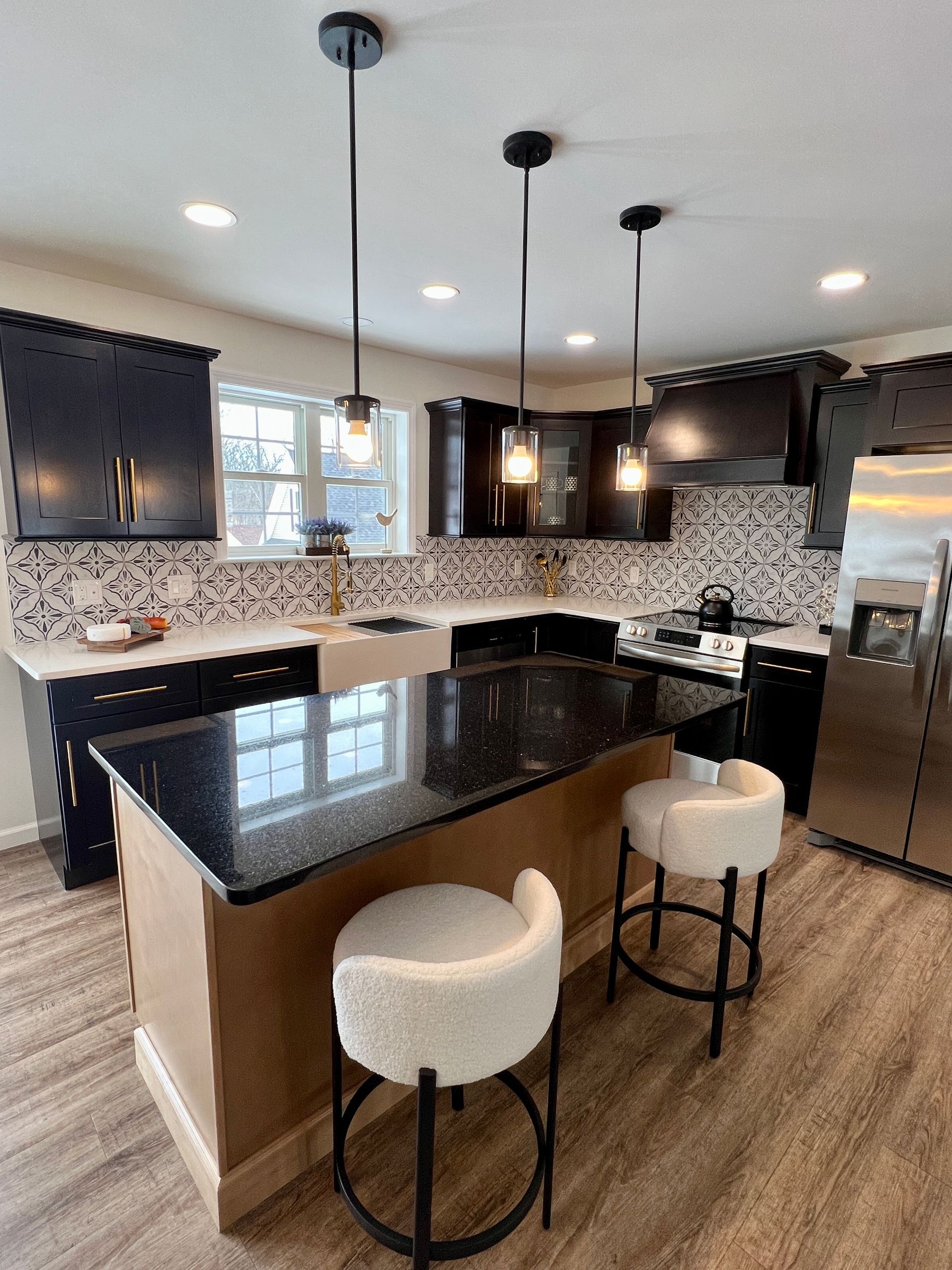 A modern kitchen featuring black cabinets, a granite-topped island with two cream-colored bar stools, and tiled backsplash.