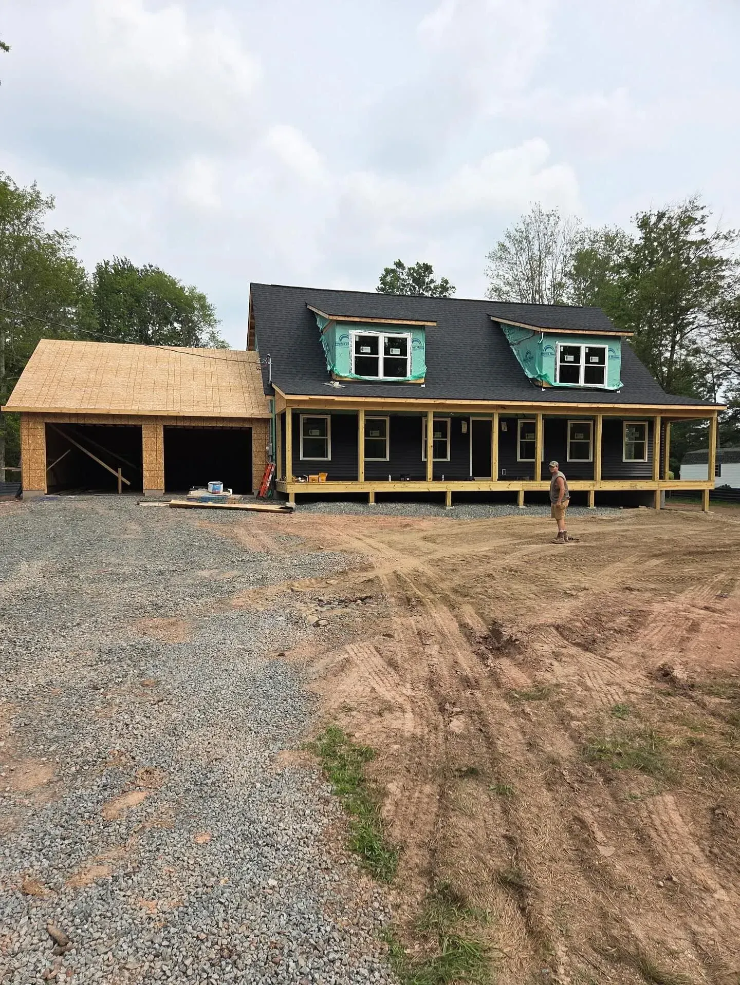 A house under construction with a gravel driveway, wooden garage framing, and dormer windows on a shingled roof.