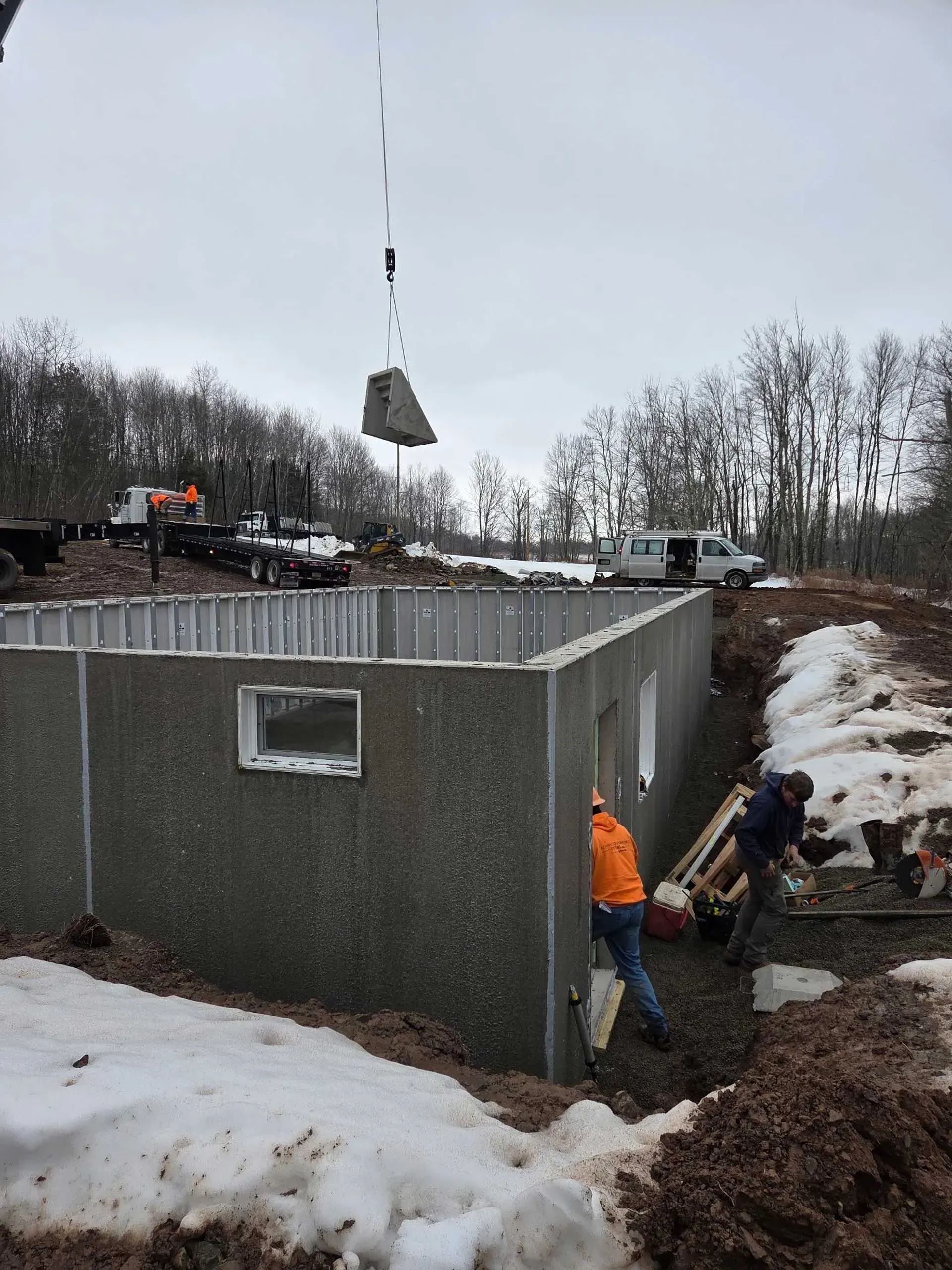 Construction workers install precast concrete foundation walls at a snowy, outdoor work site with a crane overhead.