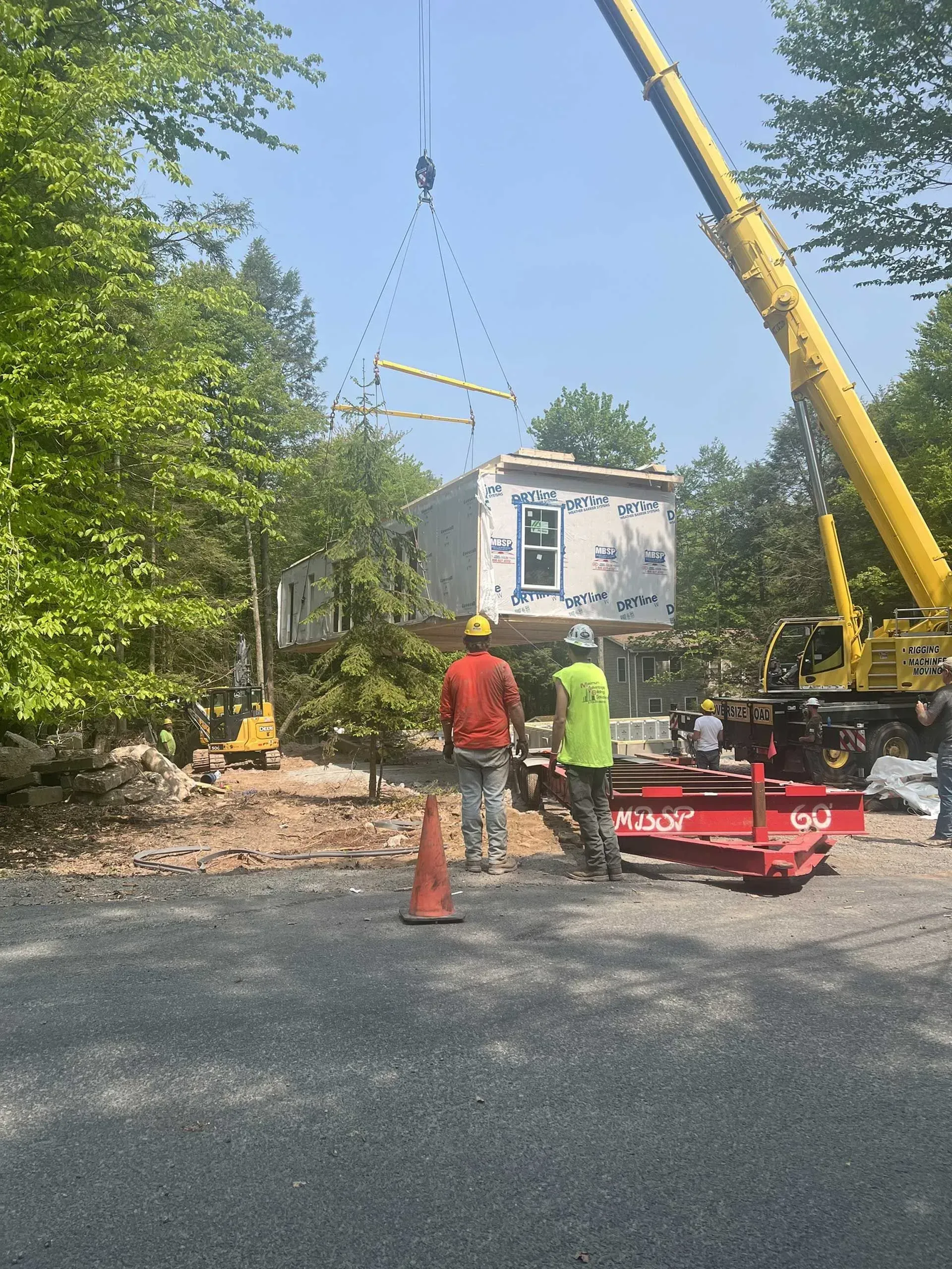A crane lifts a modular home section over construction workers on a gravel site surrounded by trees.