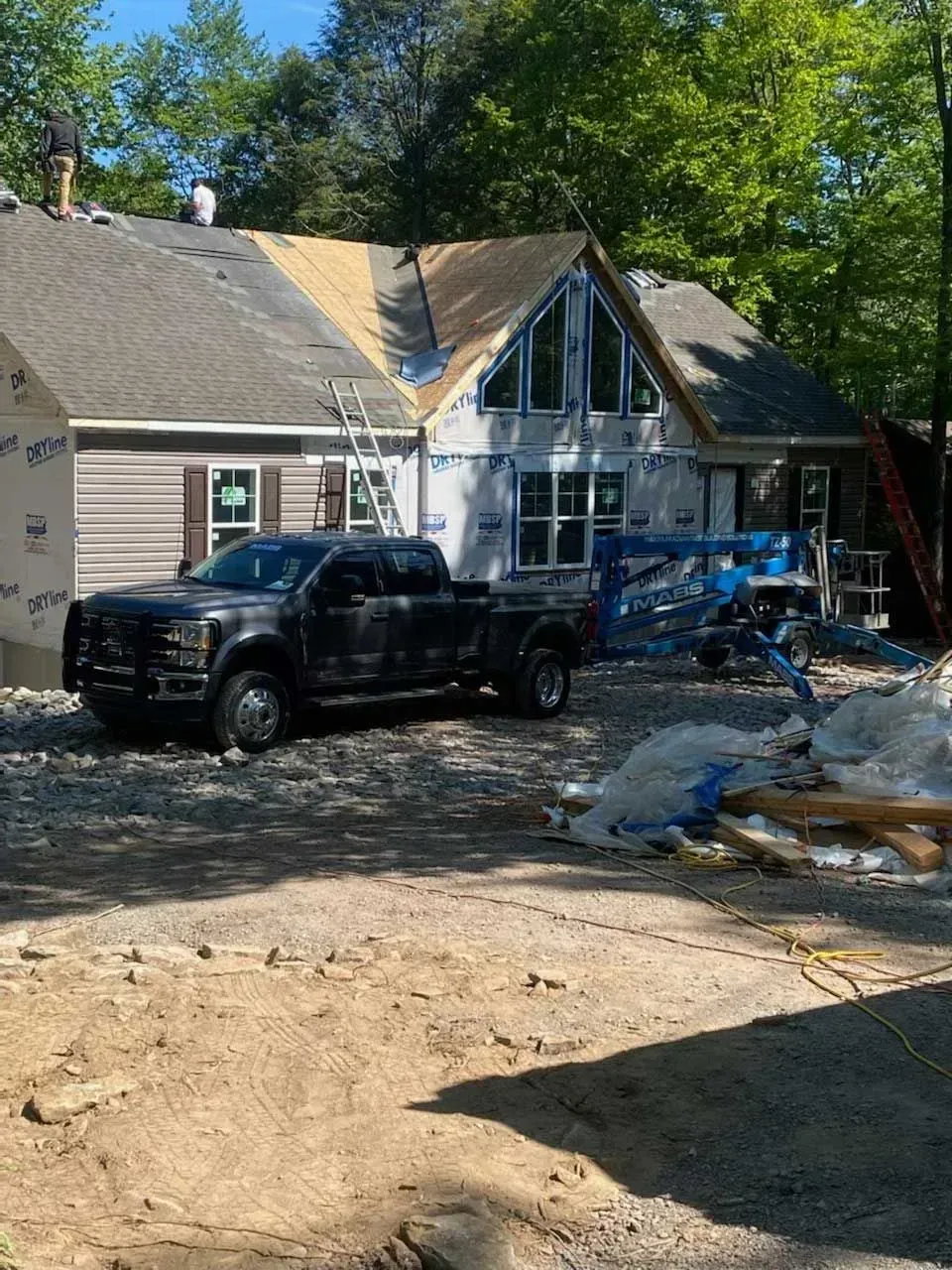 Workers repair the roof of a house under construction, with a black pickup truck and equipment parked in the yard.