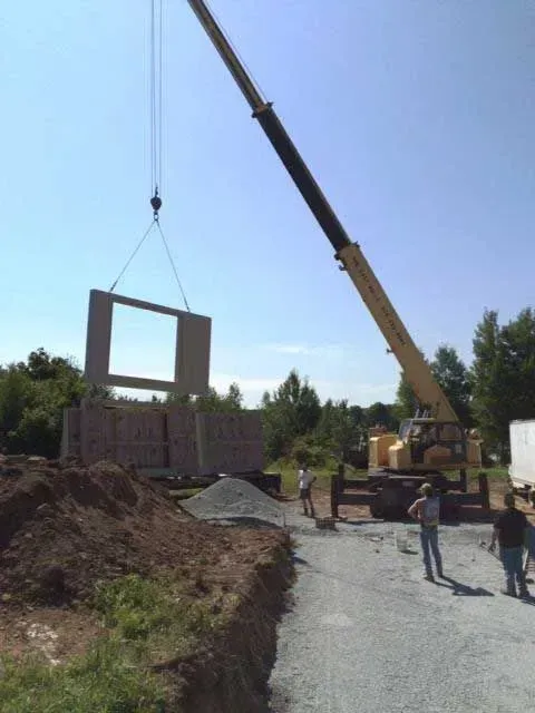 A crane lifts a large, rectangular concrete wall panel with a central window opening at a construction site.