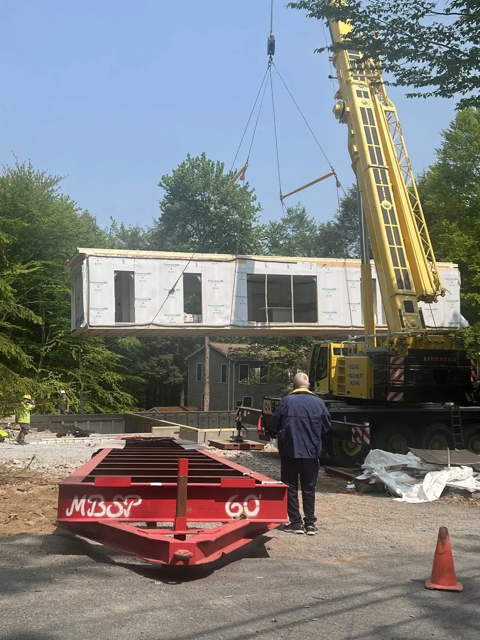 A crane lifts a modular home section above a construction site, with a trailer parked in the foreground.