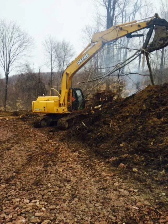 A yellow Deere excavator works on a pile of dirt in a wooded area on an overcast day.