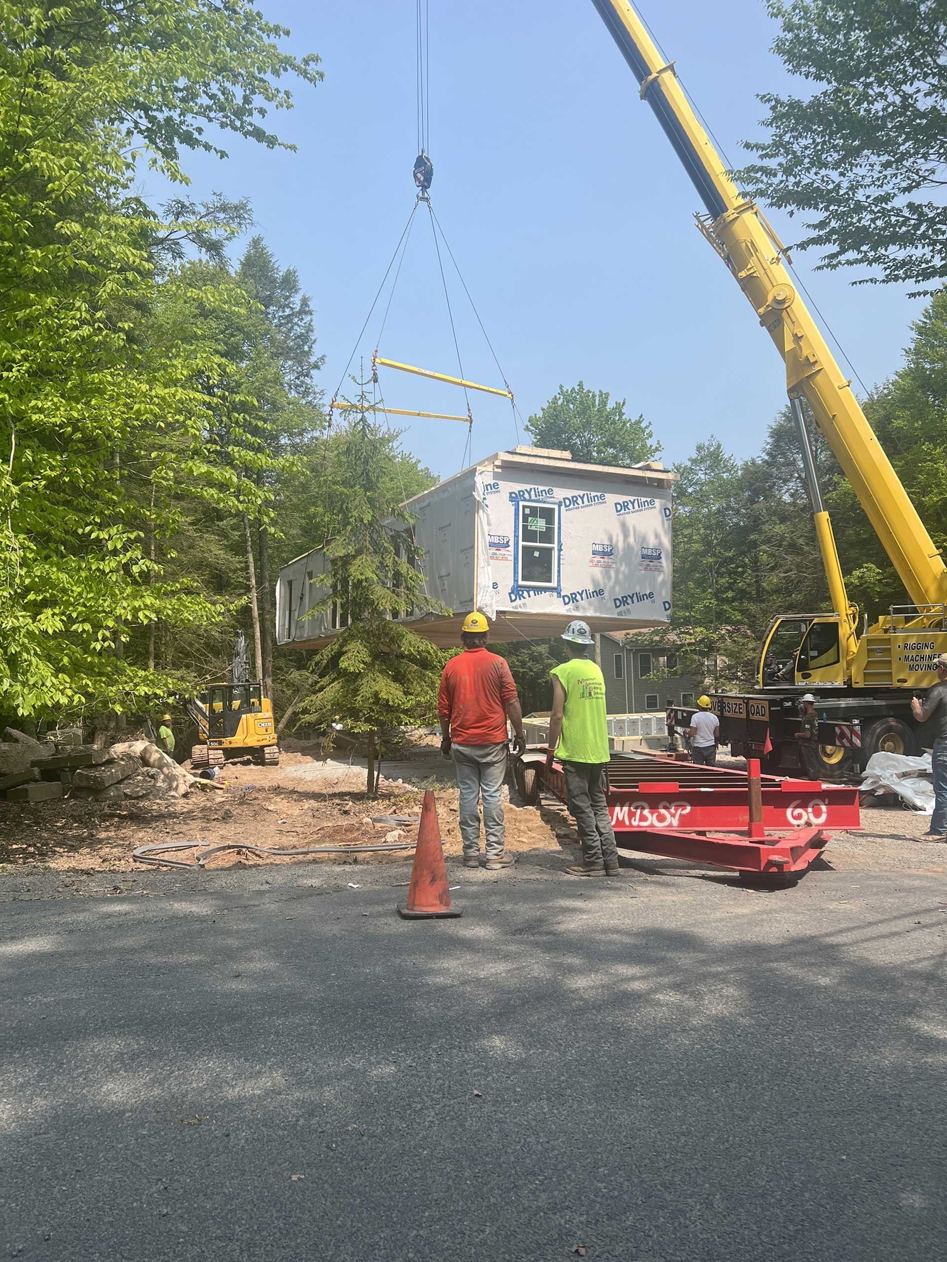 Crane lifting a modular building section; construction workers watch in a wooded area.