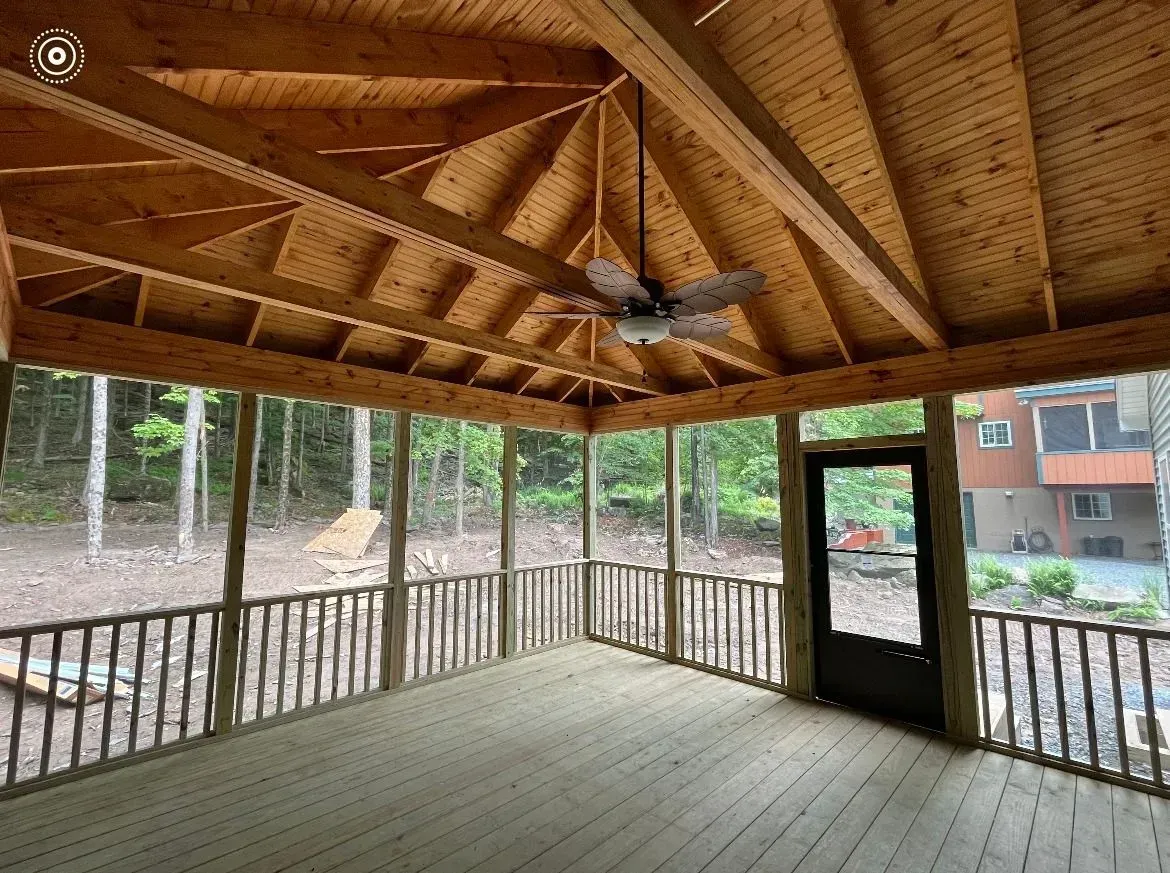 A wooden screened-in porch with a vaulted ceiling, ceiling fan, and railings overlooking a wooded outdoor area.