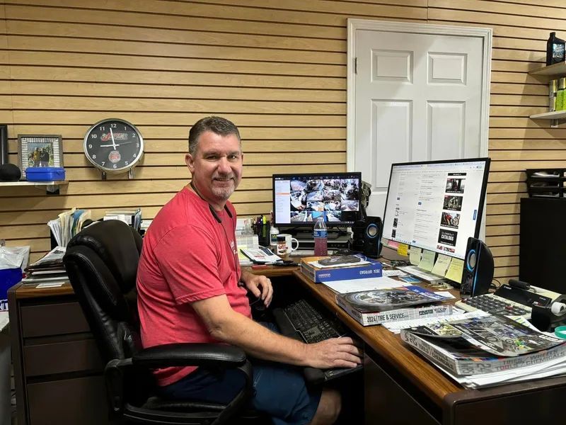 Man at a desk smiles at camera, using a computer in an office setting.