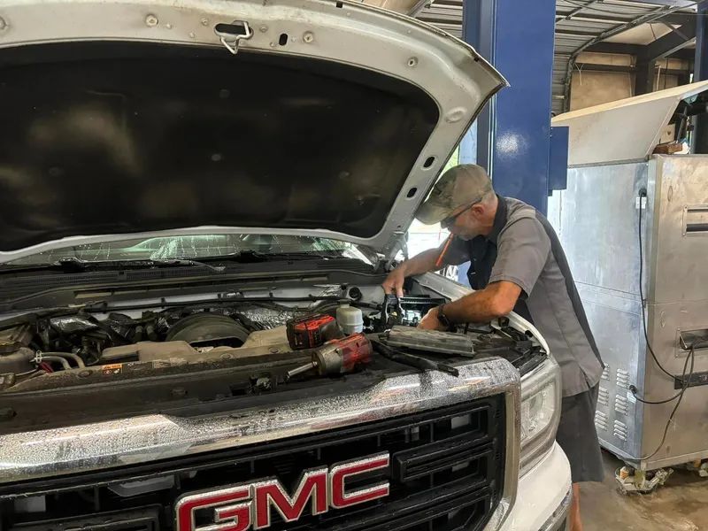 Mechanic working on a white GMC truck with hood open in a shop. Tools are nearby.