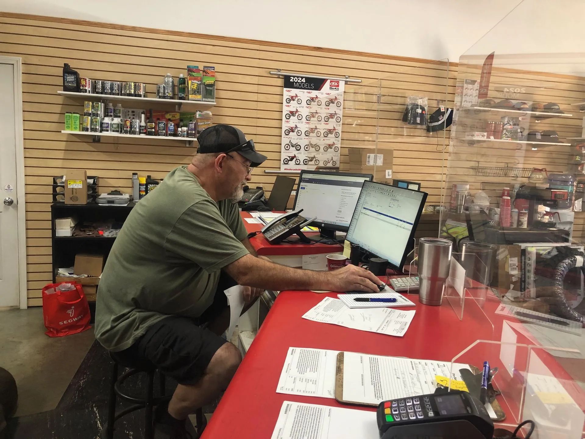 Man in a baseball cap works at a red counter, looking at a computer screen. Motorcycle parts on shelves in the background.