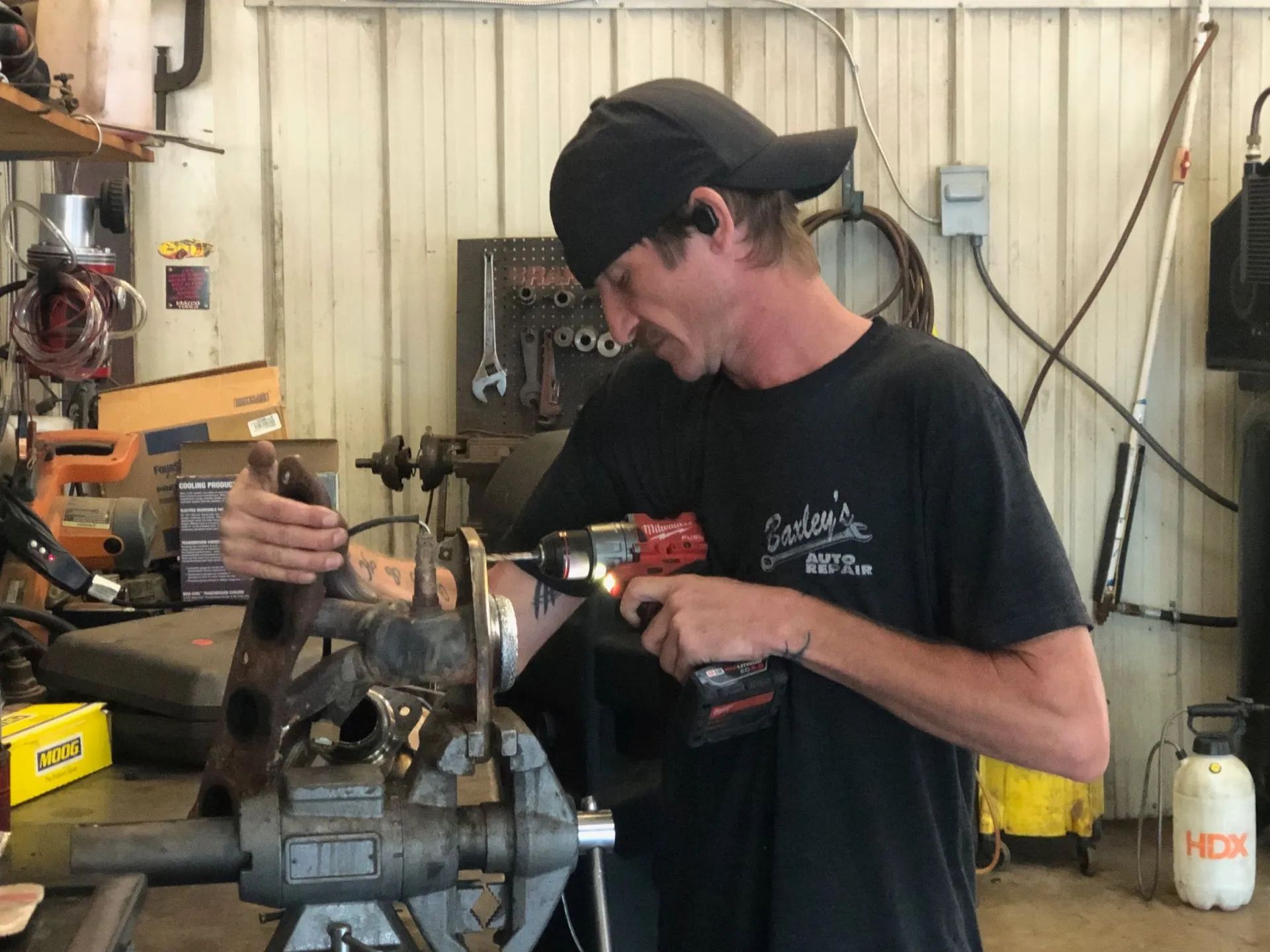 Man in a workshop using a drill on a metal object held in a vise. He wears a black cap and shirt.