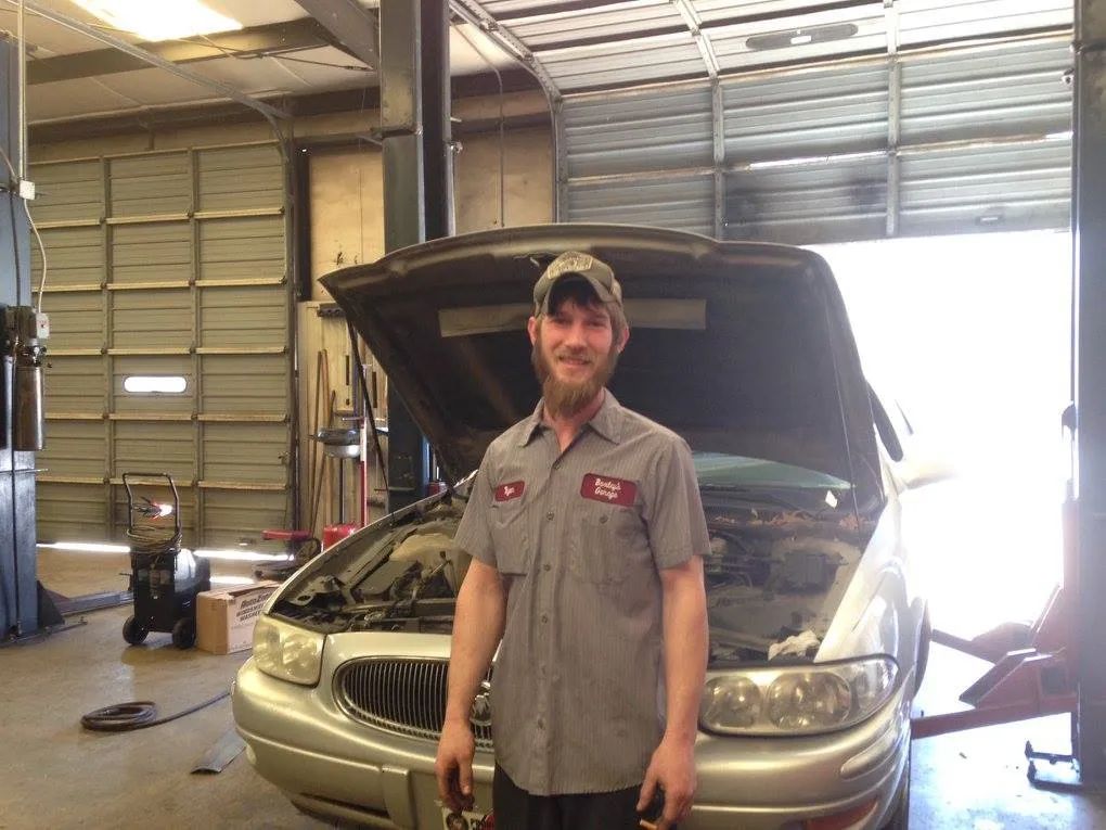 Mechanic in grey shirt and cap stands in front of a car with an open hood inside a garage.