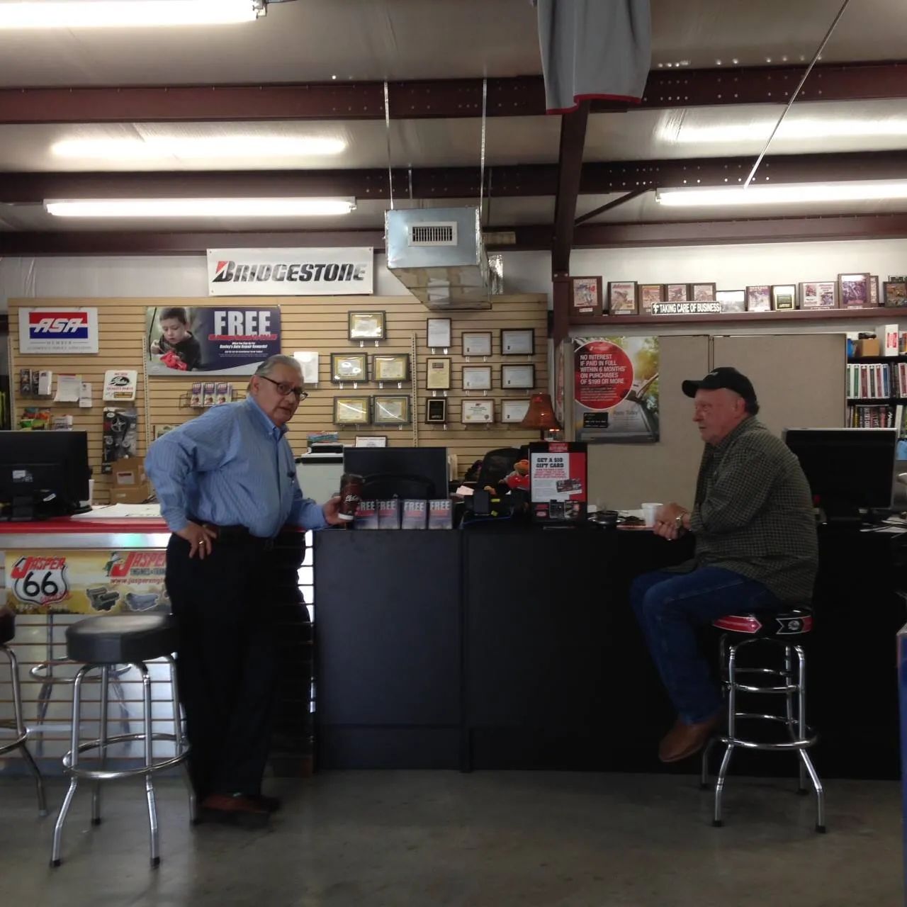 Two men in an auto parts store. One stands, the other sits. Counter with awards behind them.