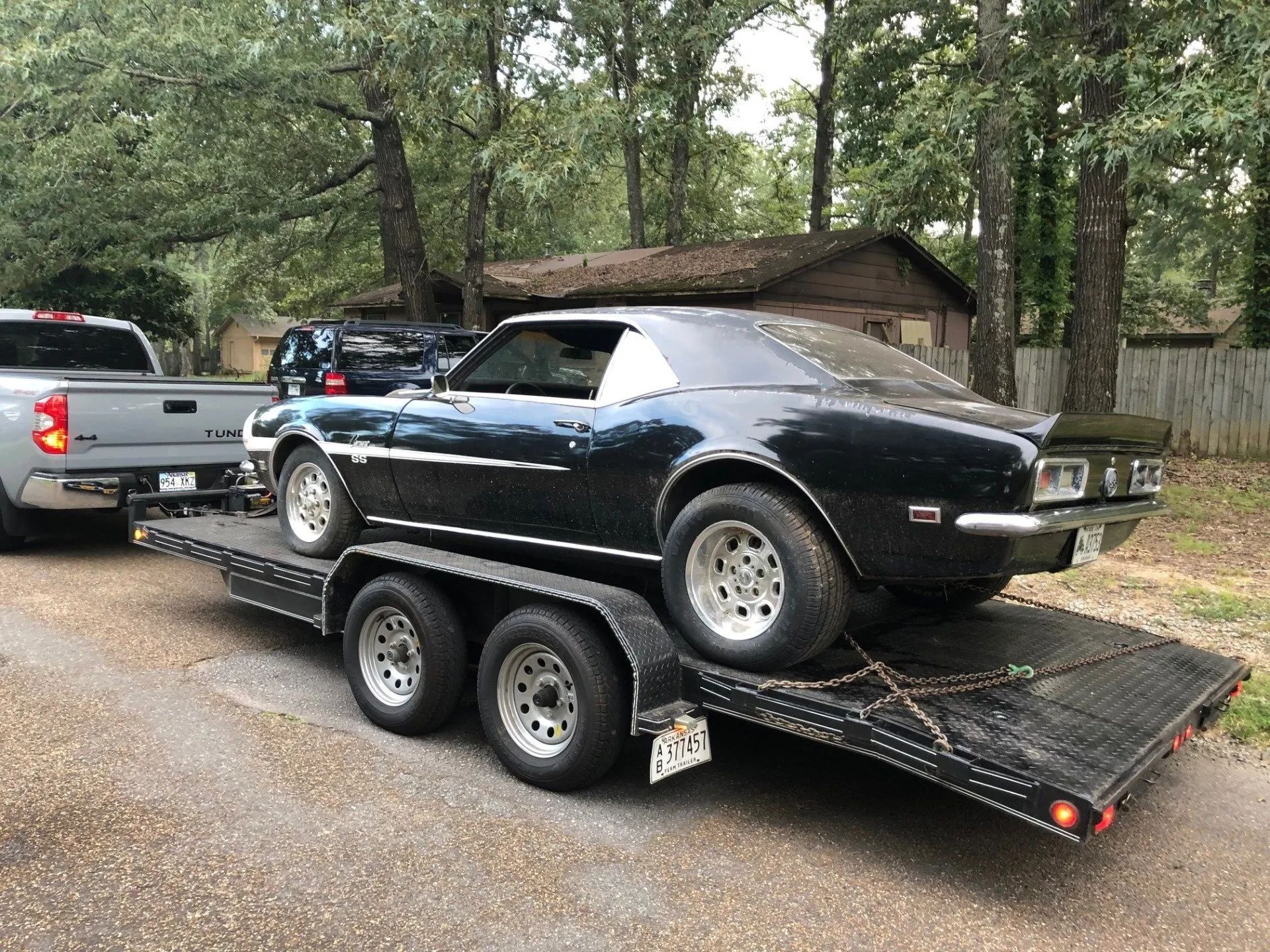Black classic car on a trailer being towed by a gray pickup truck in a residential area.
