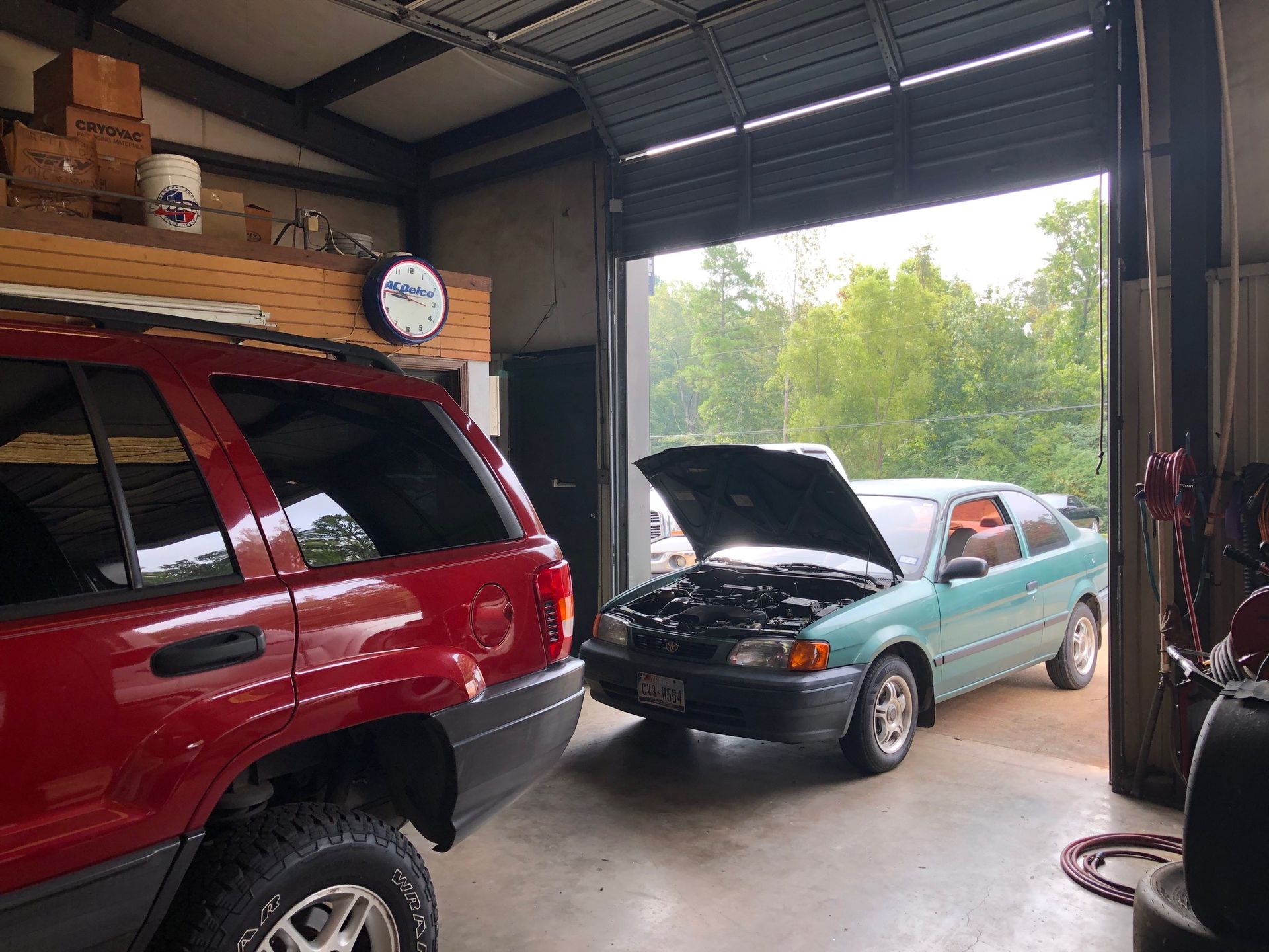 Red Jeep and teal car in a garage with open door, view of trees. Car hoods are up.