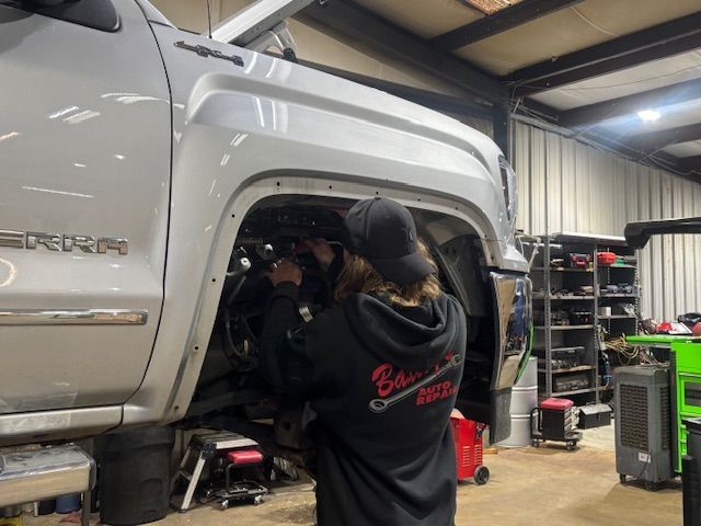 A person working on a silver truck in a garage; they are wearing a black hoodie and cap.