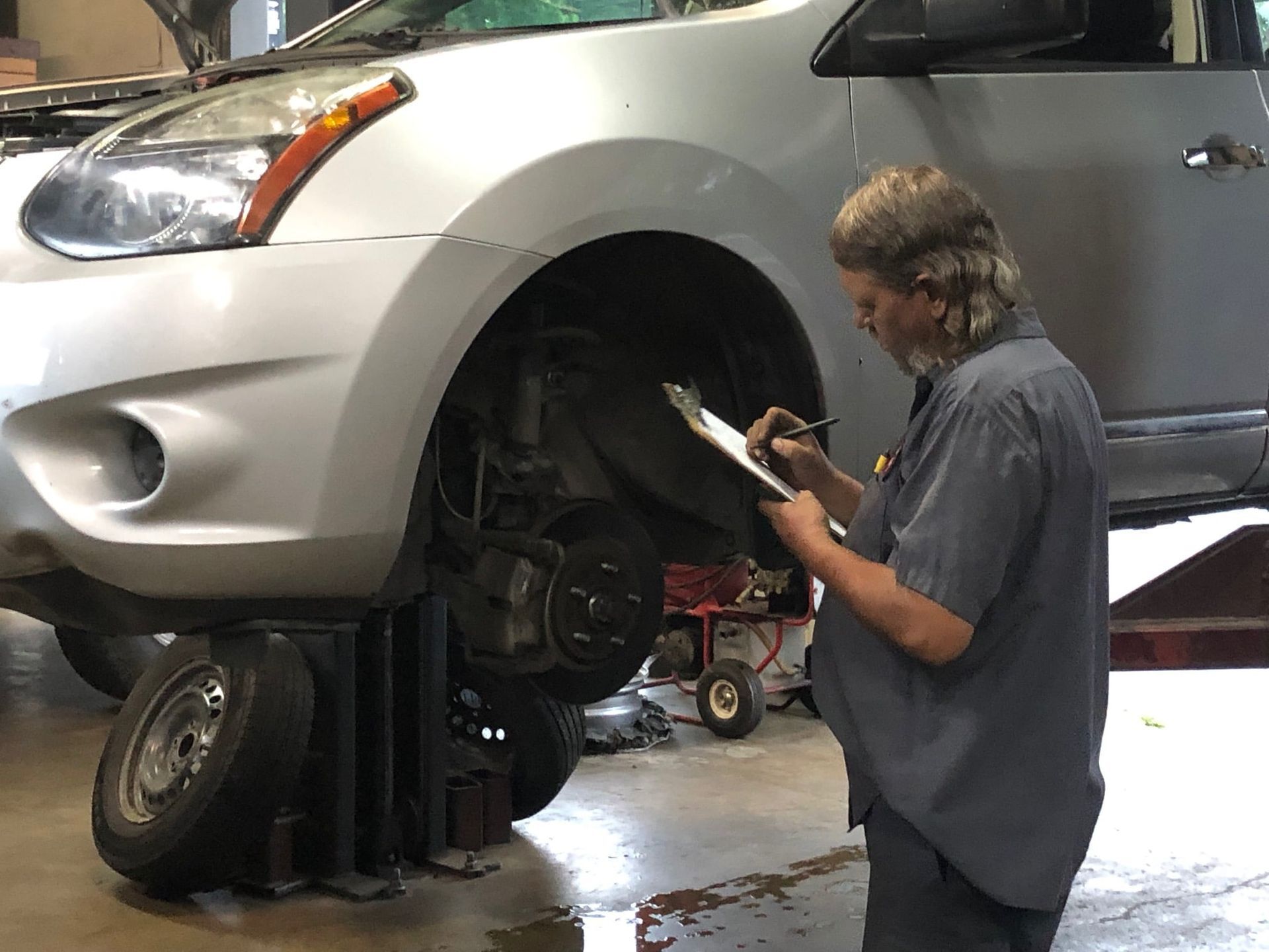 Mechanic in a gray uniform inspecting a car's wheel area while holding a clipboard. Car is raised on a lift.