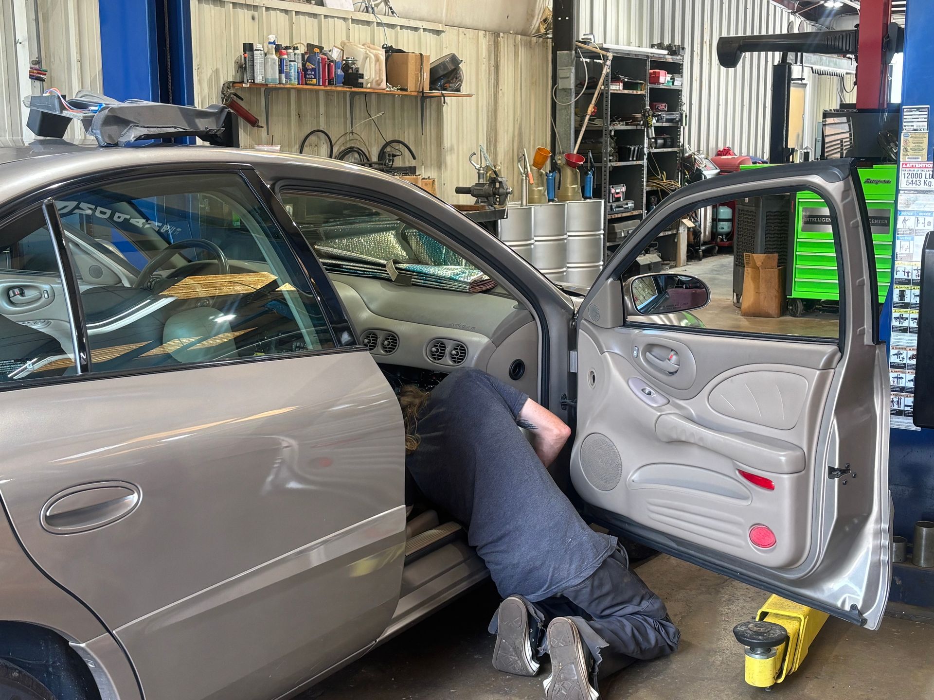 Mechanic working inside a gray car at an auto shop, with the driver door open.