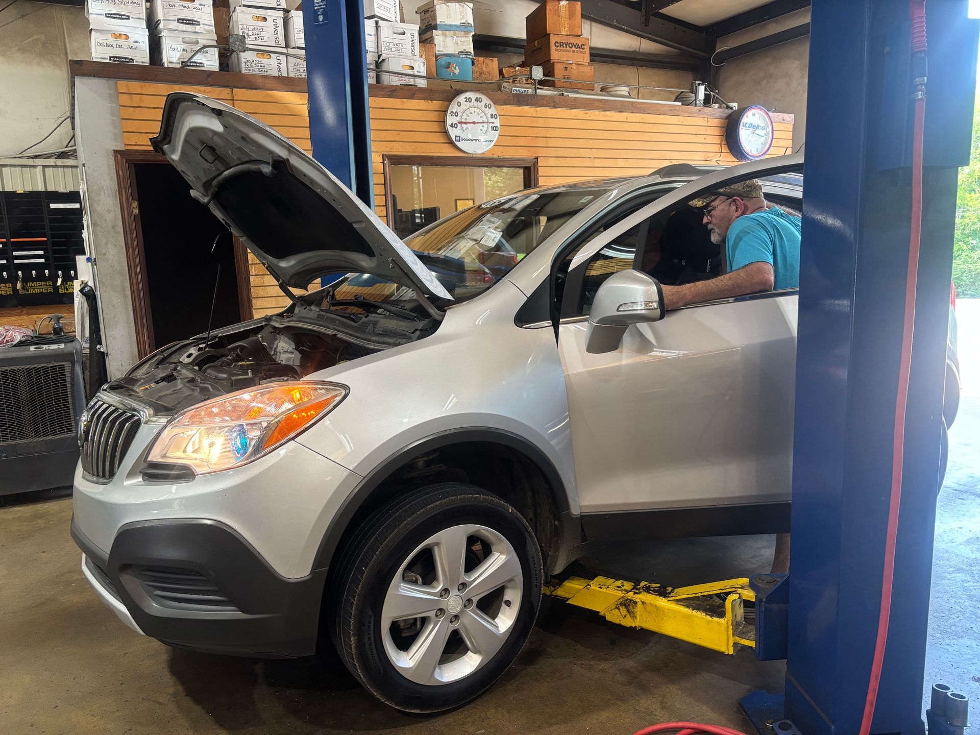 Silver SUV on a car lift in an auto repair shop, hood open and a mechanic working inside.