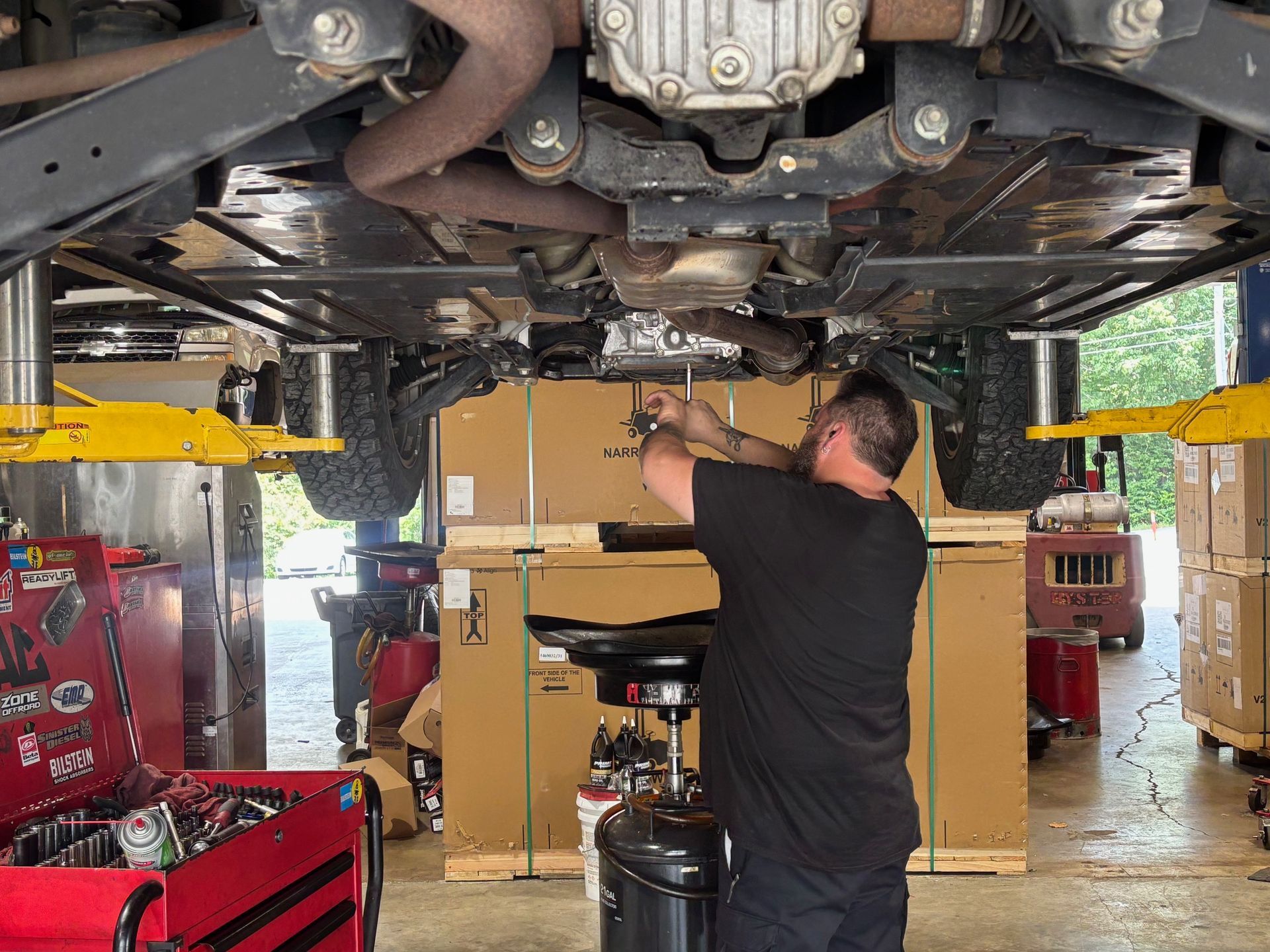 Mechanic working under a raised vehicle in a garage, reaching up with tools among car parts and boxes
