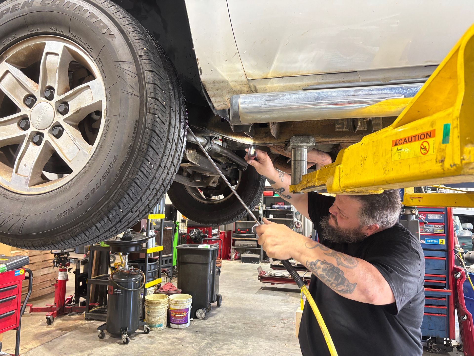 Mechanic working under a lifted silver car in a garage, using a tool near the rear wheel.
