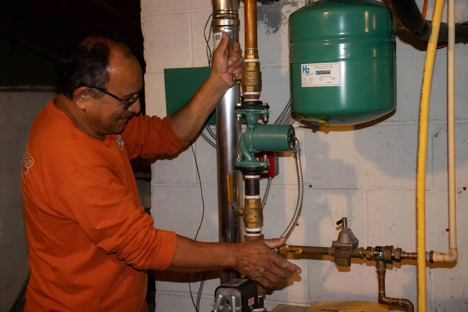 Man in orange shirt working on pipes in a basement, near a green tank and other plumbing.