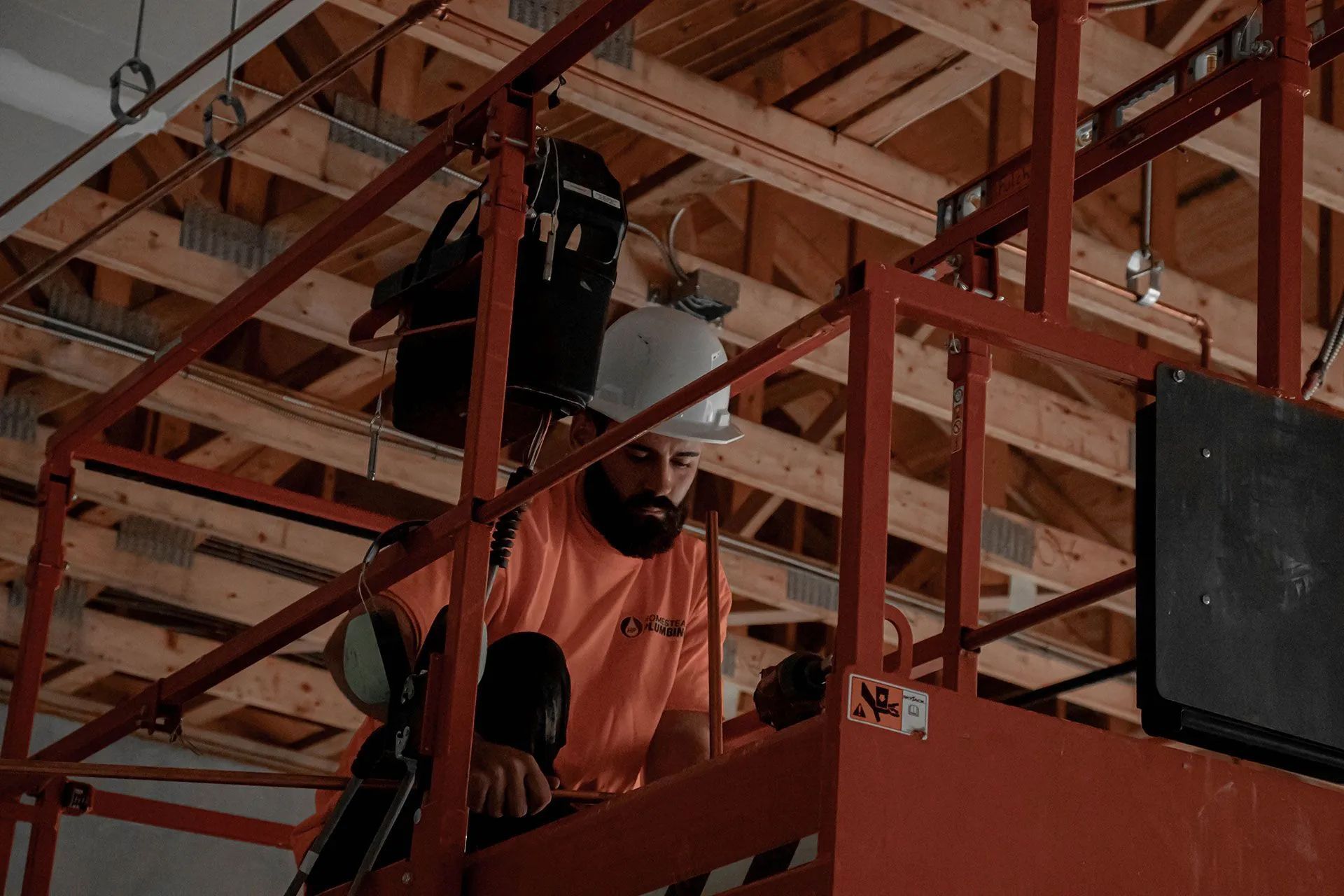 Construction worker in an orange shirt and white hard hat works on ceiling from a red lift.