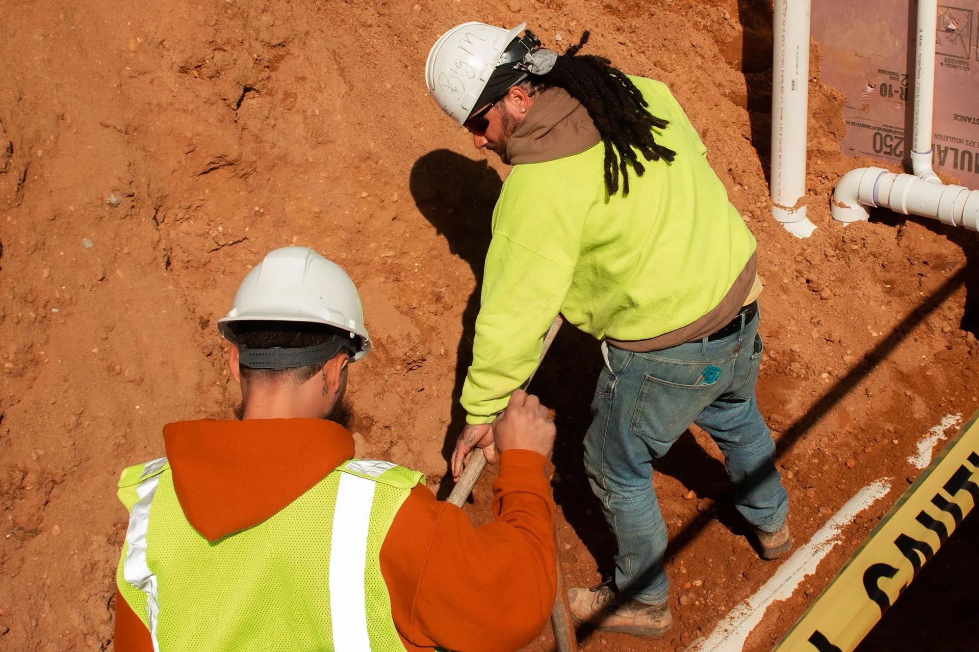 Two construction workers digging in an excavation, one wearing a yellow safety vest and the other a white hard hat.