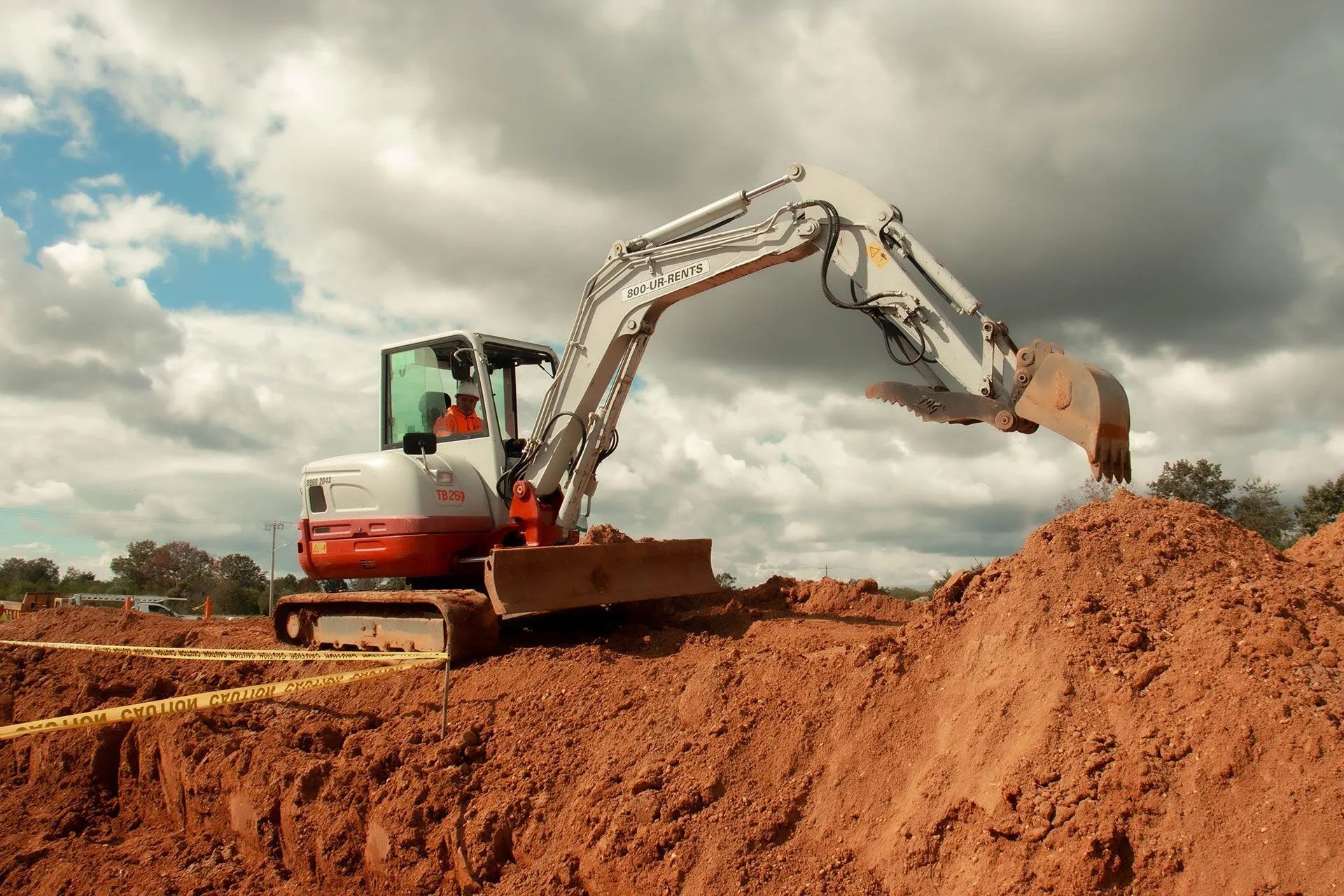 Small excavator digging in reddish soil under a cloudy sky.
