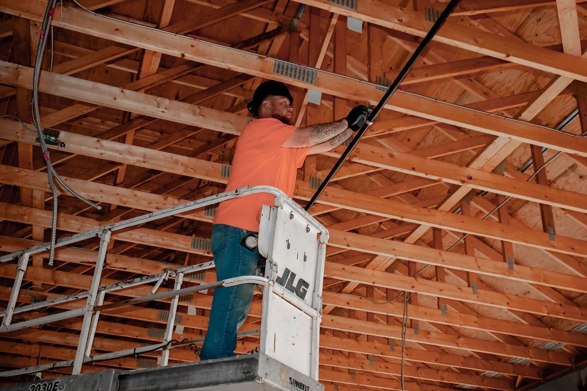 Construction worker on lift, reaching up to work on overhead black pipe within wooden roof structure.