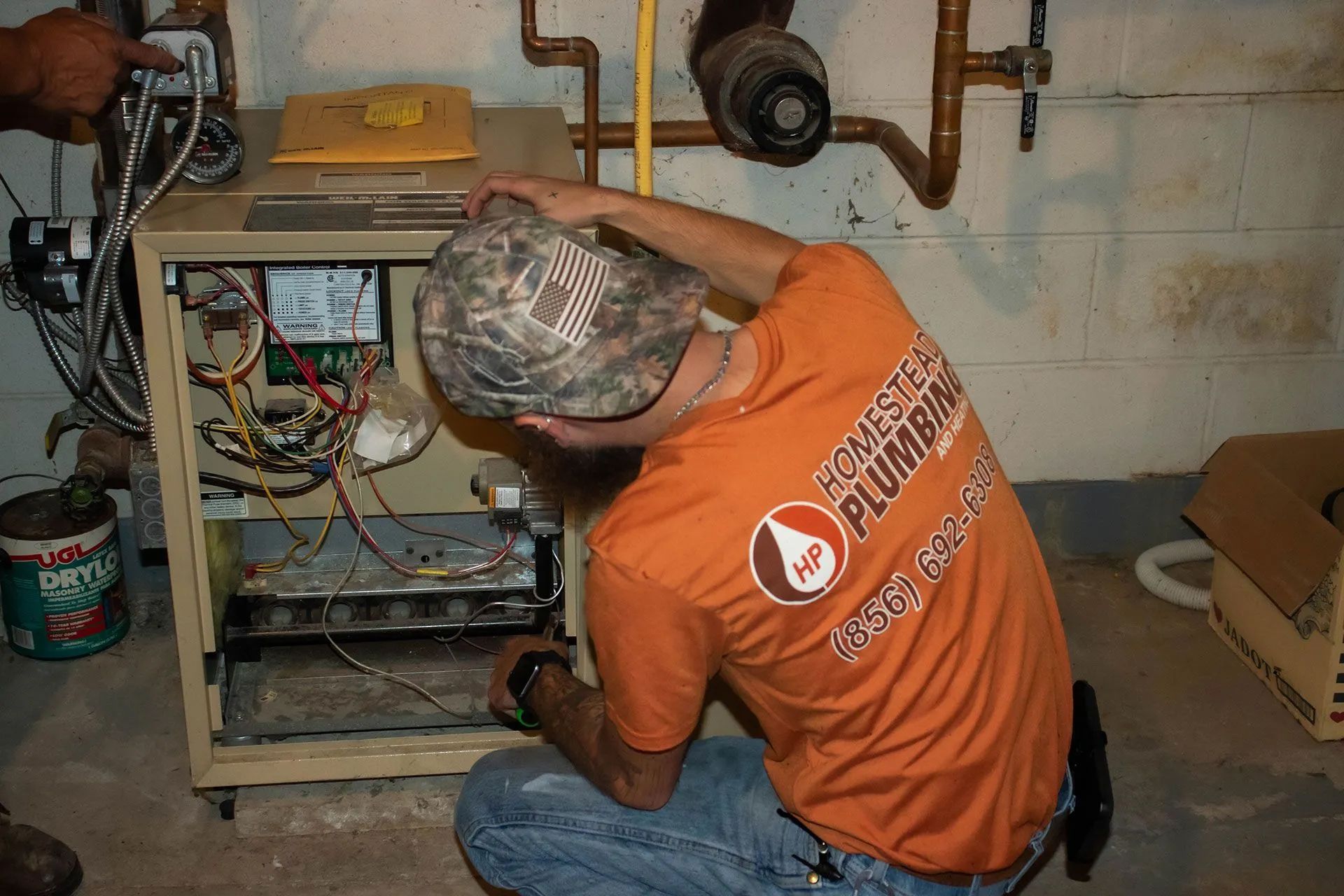 Plumber in an orange shirt repairs furnace in a basement, checking wires.