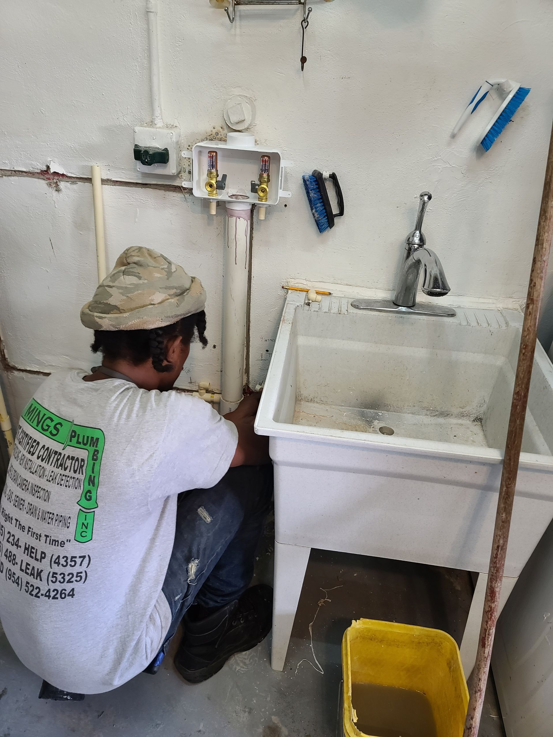 Plumber connecting pipes near a utility sink in a utility room. Yellow bucket is on floor.