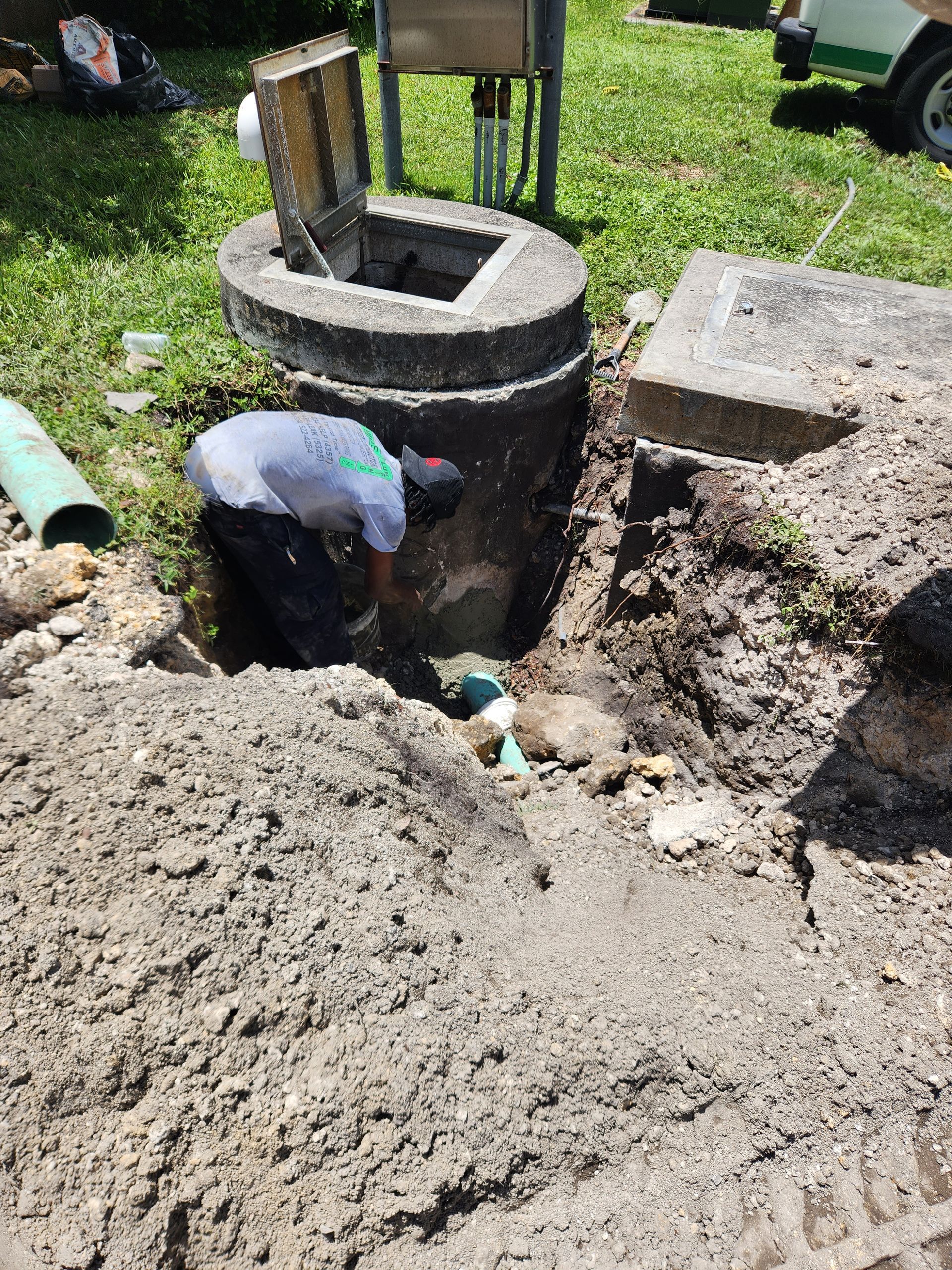 Man working on plumbing near an open concrete access point in a yard. Soil surrounds the hole.