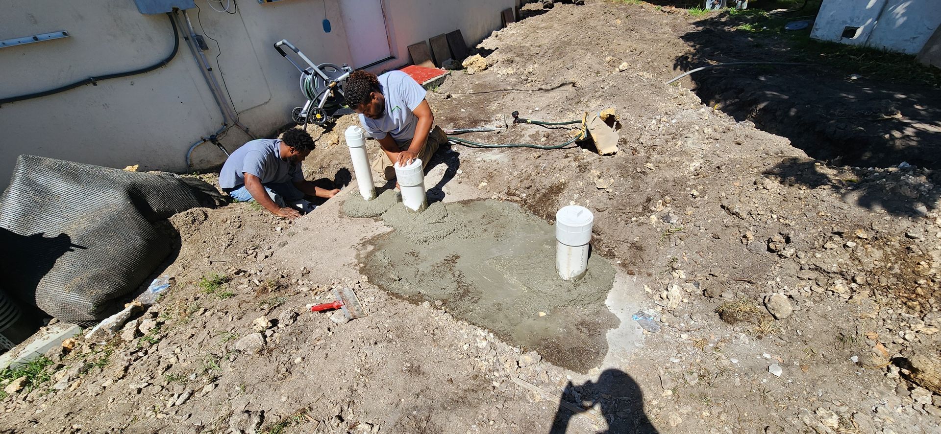 Two people working on a concrete project outdoors. One kneels by concrete base; another works on piping.