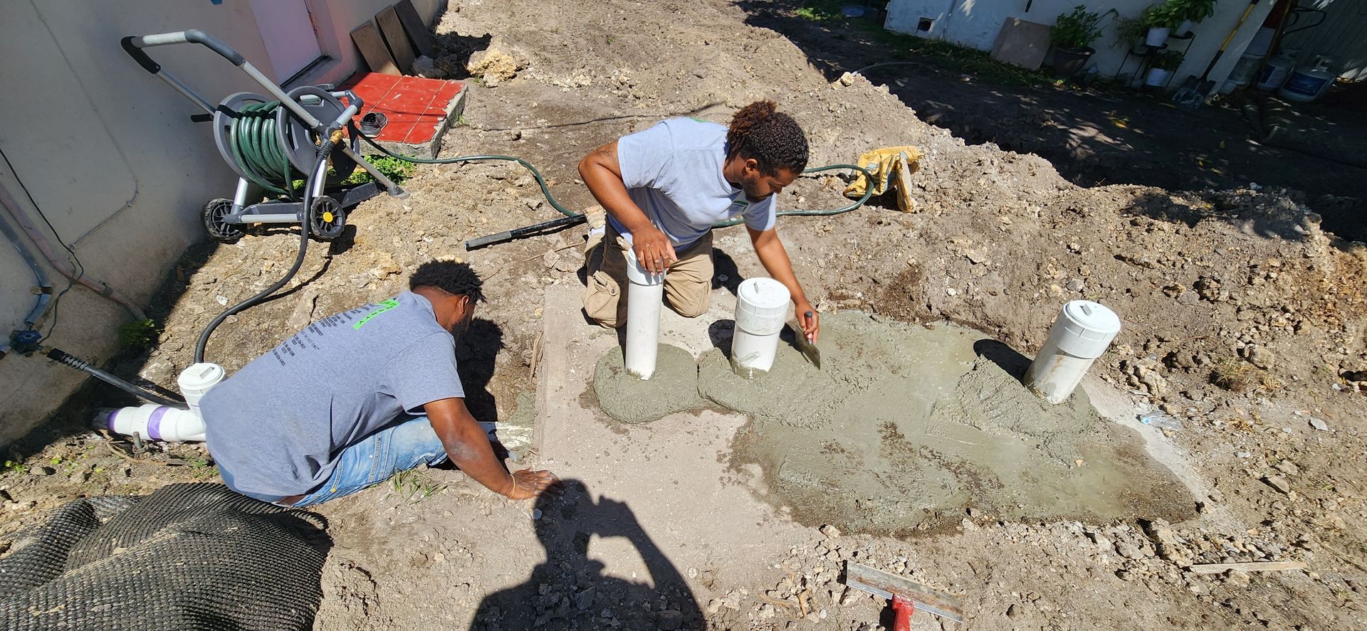 Two people working on a concrete project outdoors. One is holding a tool, another is working with pipes.