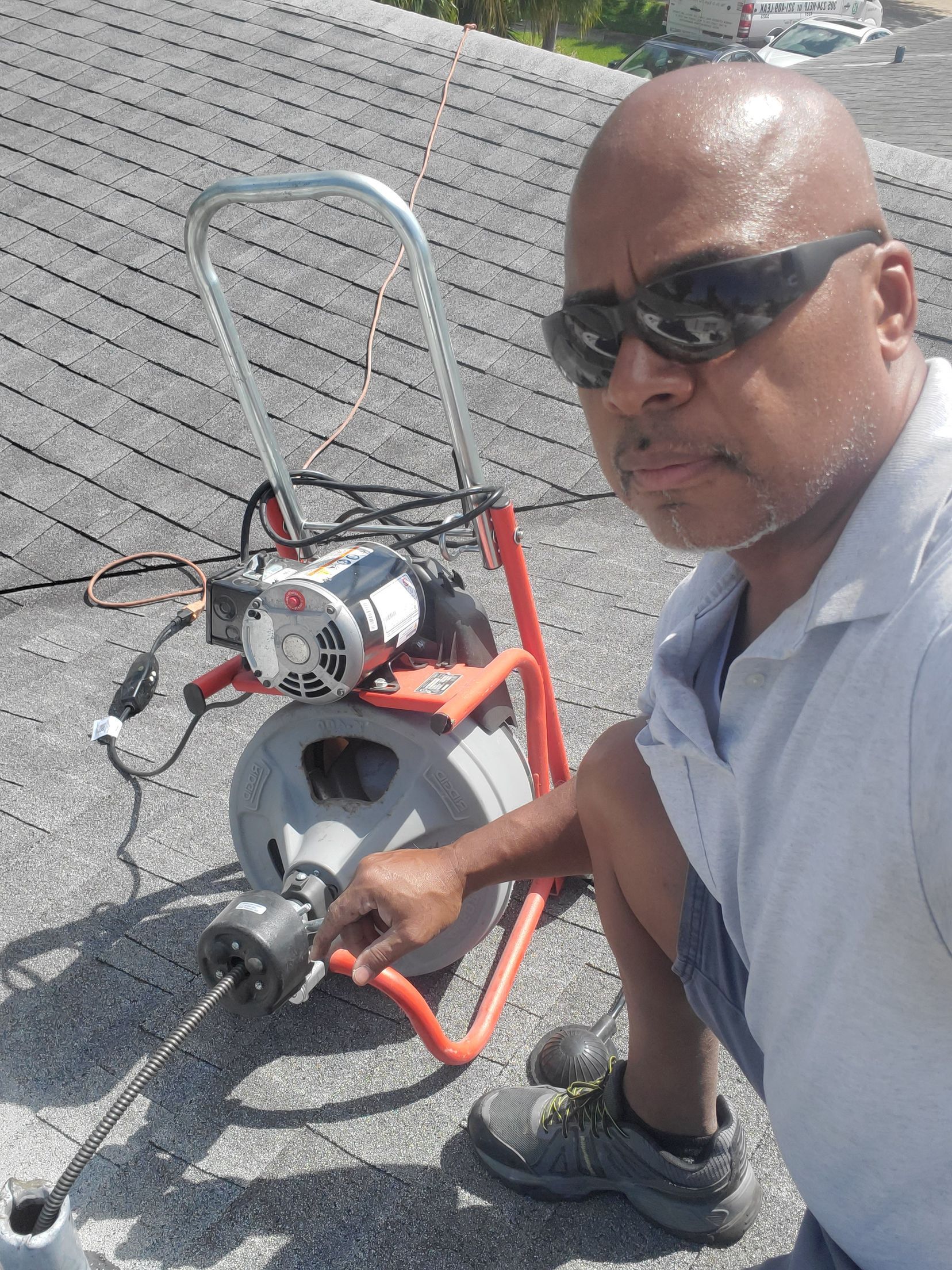 Man operating a drain snake on a rooftop. He's wearing sunglasses and work boots. A red and silver machine is behind him.