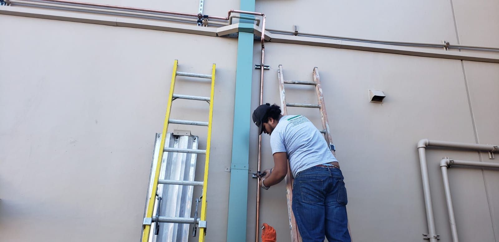 A person in blue jeans and a light blue shirt works on pipes against a building wall, two ladders next to them.