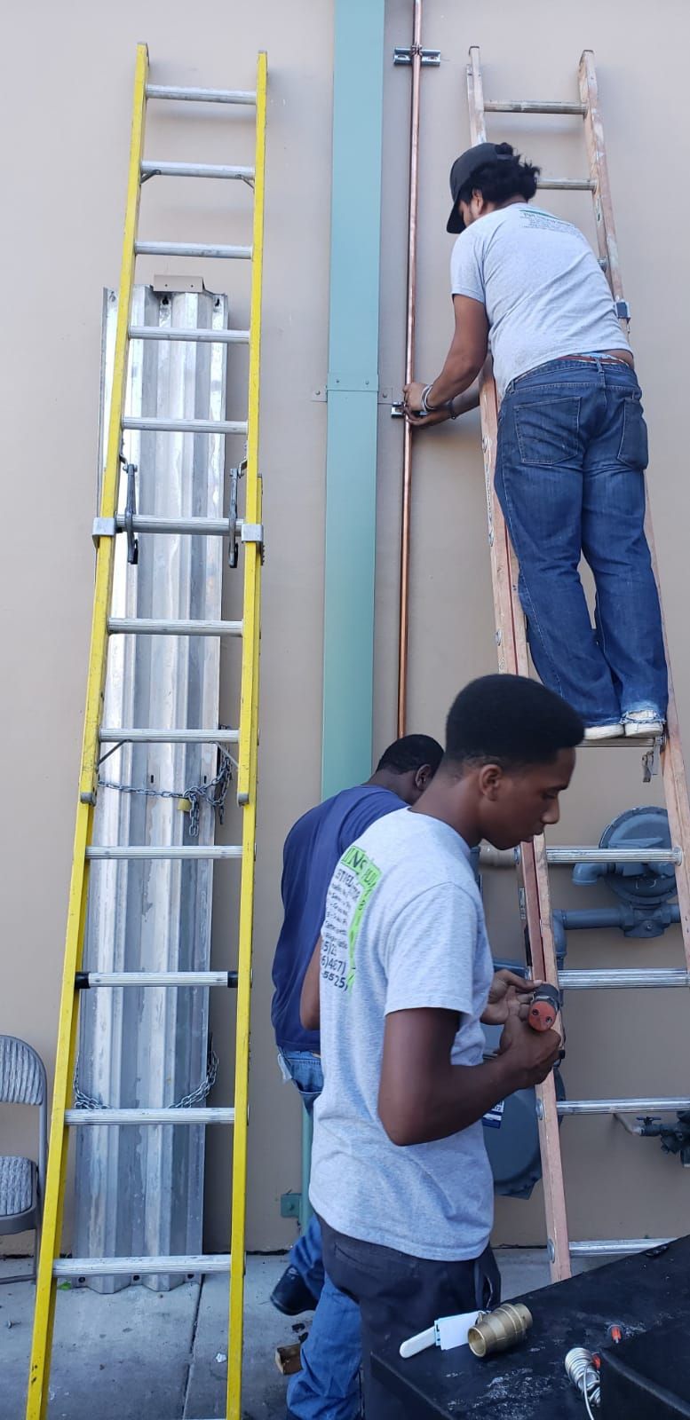 Three people working on a structure outdoors using ladders. One person is on a ladder working on copper piping.