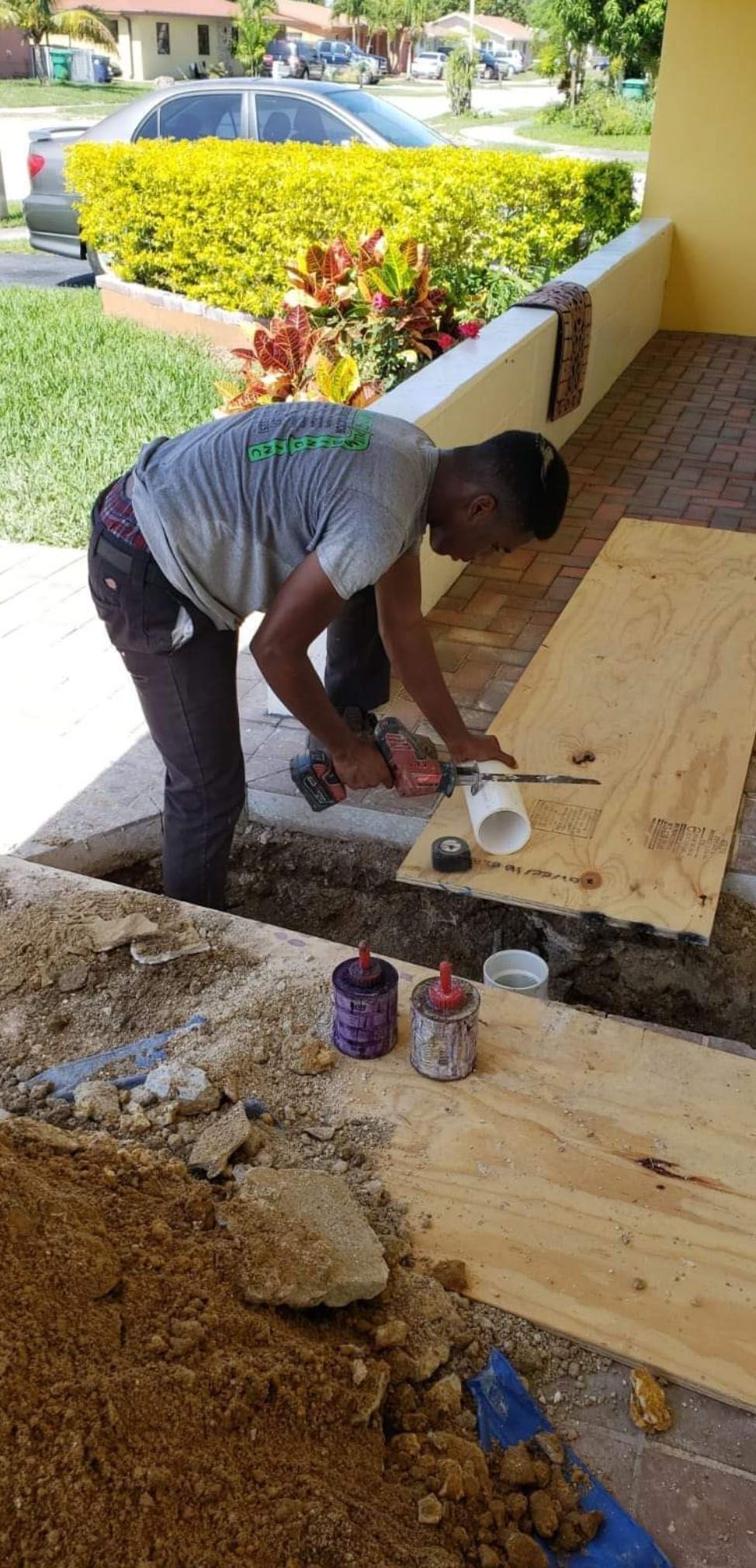 A person working on plumbing outdoors, holding a tool near white pipe, next to wood and soil.