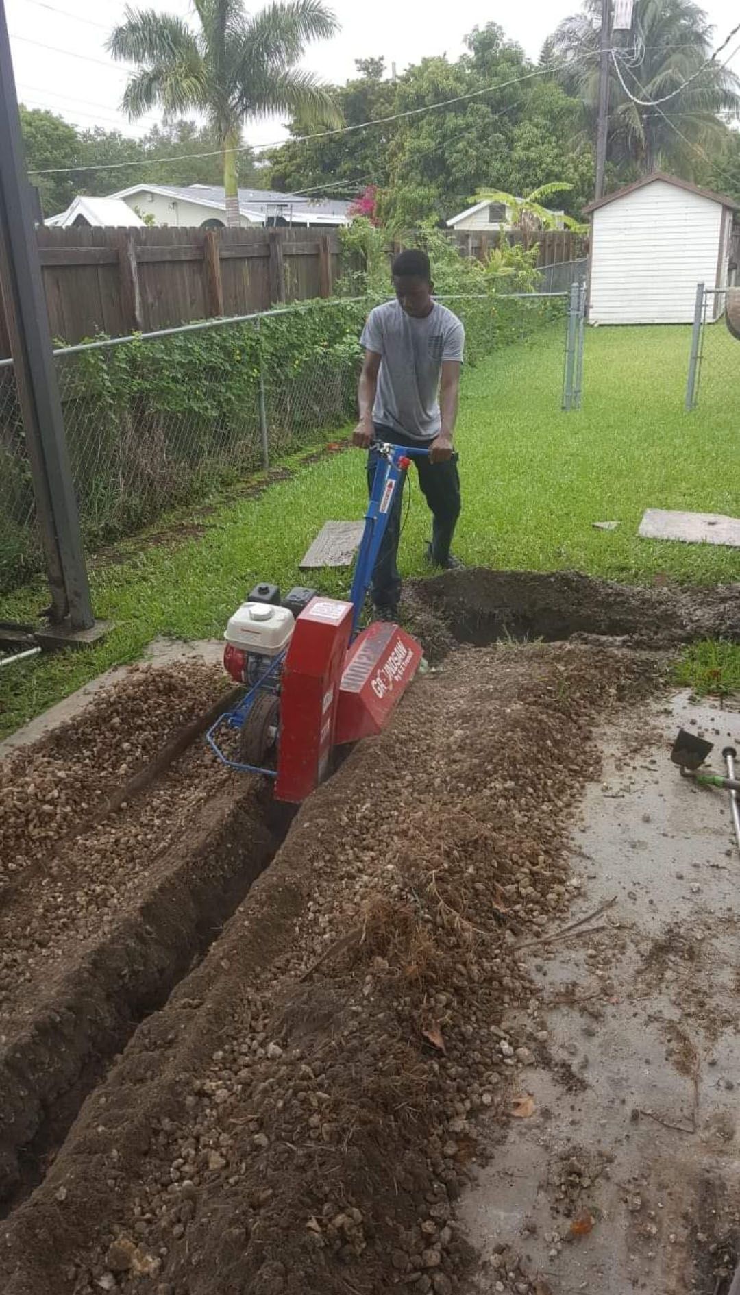 Man using a trencher in a yard, digging a trench in the soil next to a concrete surface.