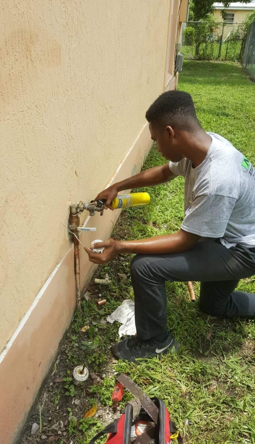 Plumber kneeling, soldering pipe near tan building exterior. Green grass visible.