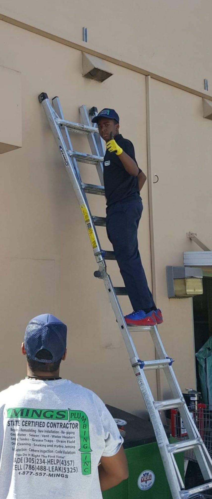 Two men working near a wall. One is on a ladder, and the other looks on.