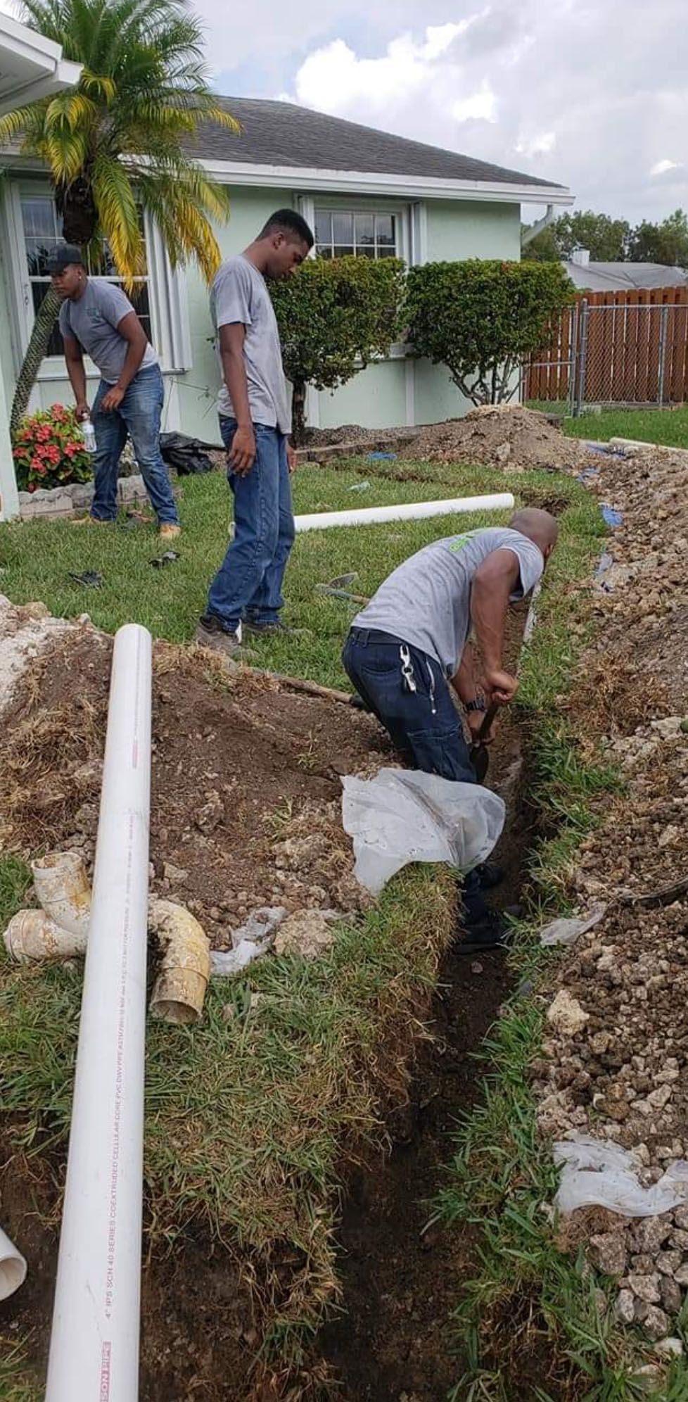 Three people digging a trench in a grassy yard, near a house with a white pipe running alongside.