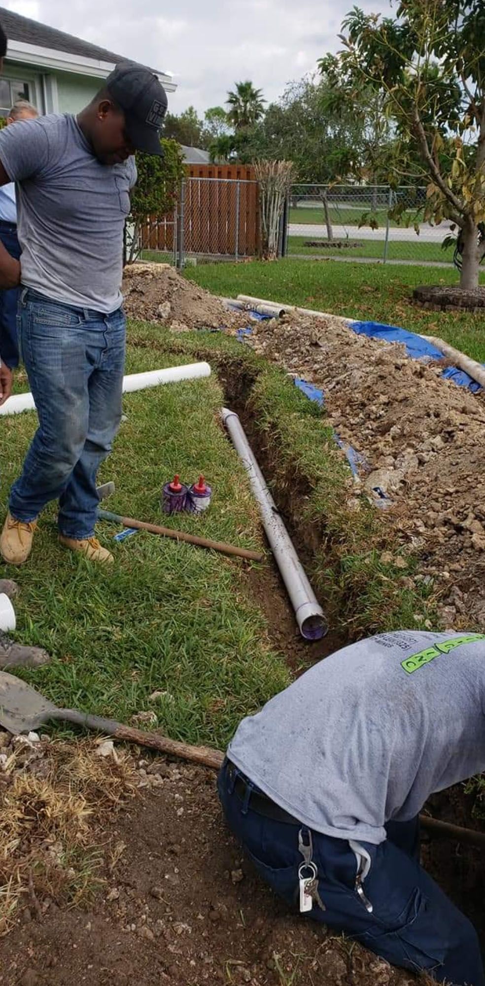 Workers install pipes in a ditch dug in a grassy yard. One kneels. Two others stand nearby.