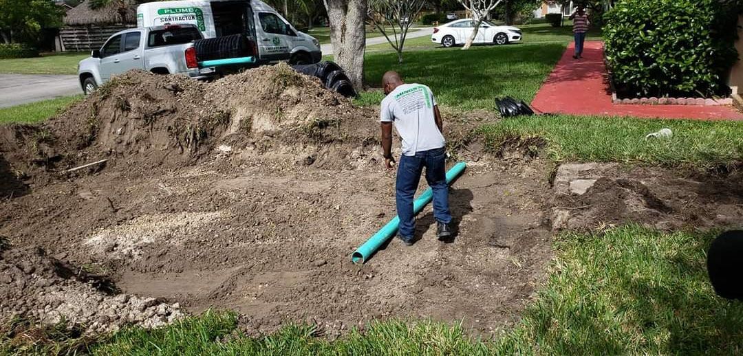 A man in a white shirt and blue jeans working on a ditch in a yard, with piping.