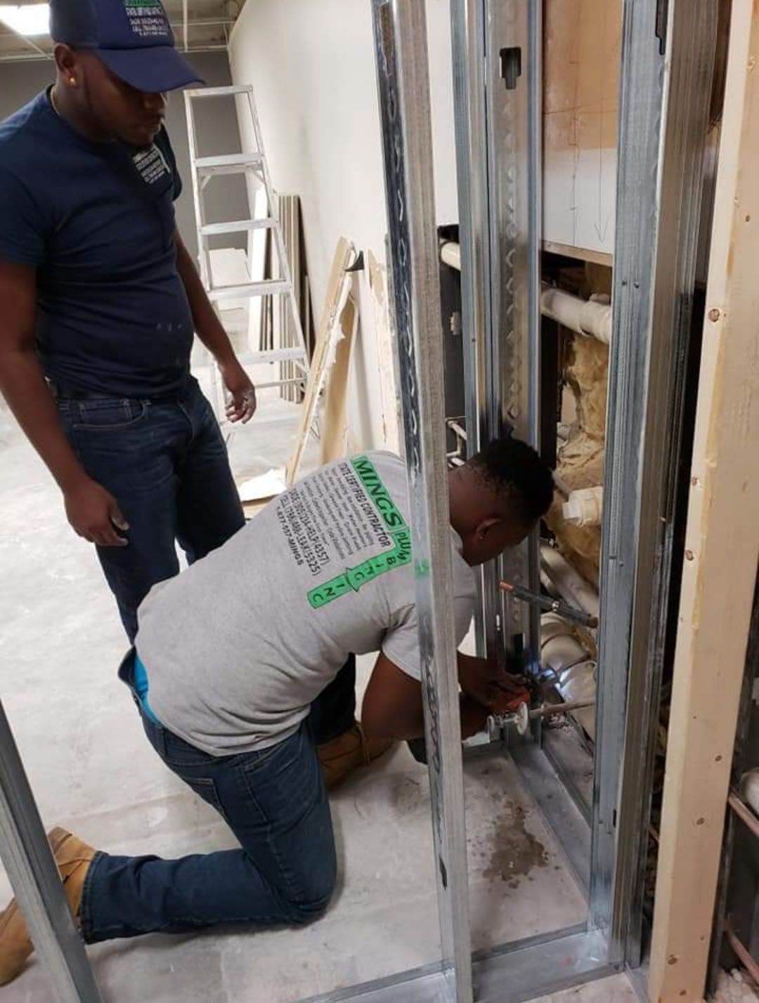 Two workers installing plumbing pipes within a metal stud frame in a construction site.