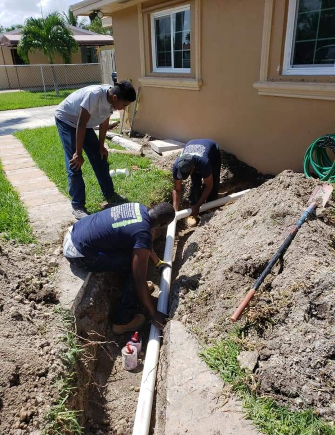 Three people installing a white pipe in a trench beside a beige building.