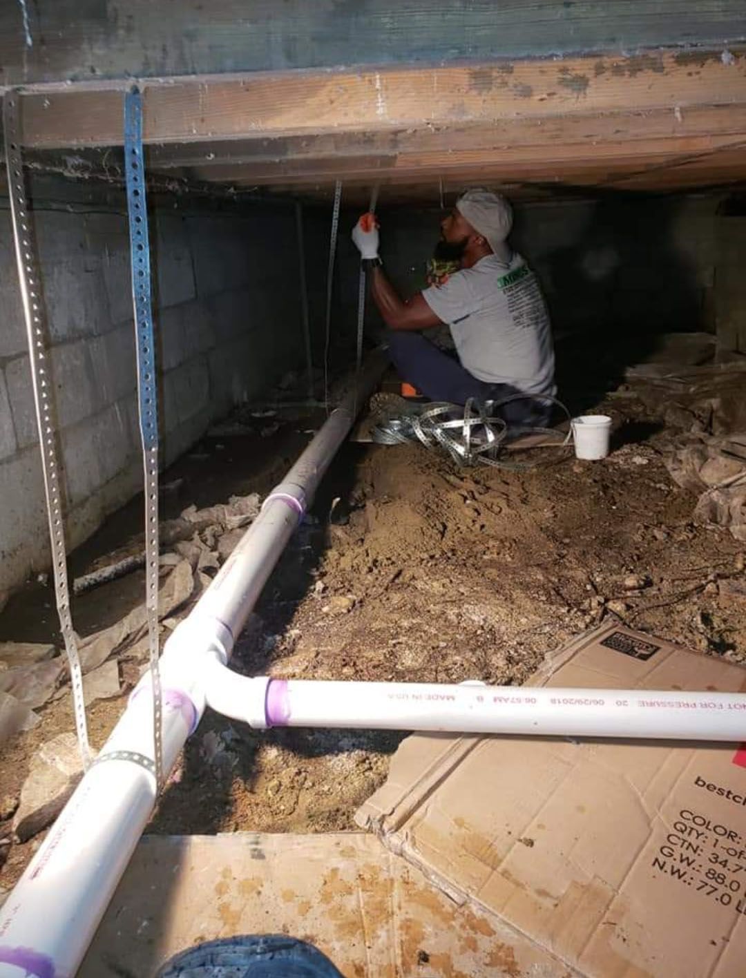 Plumber working on PVC pipes in a crawlspace. White and purple pipes against dark background.
