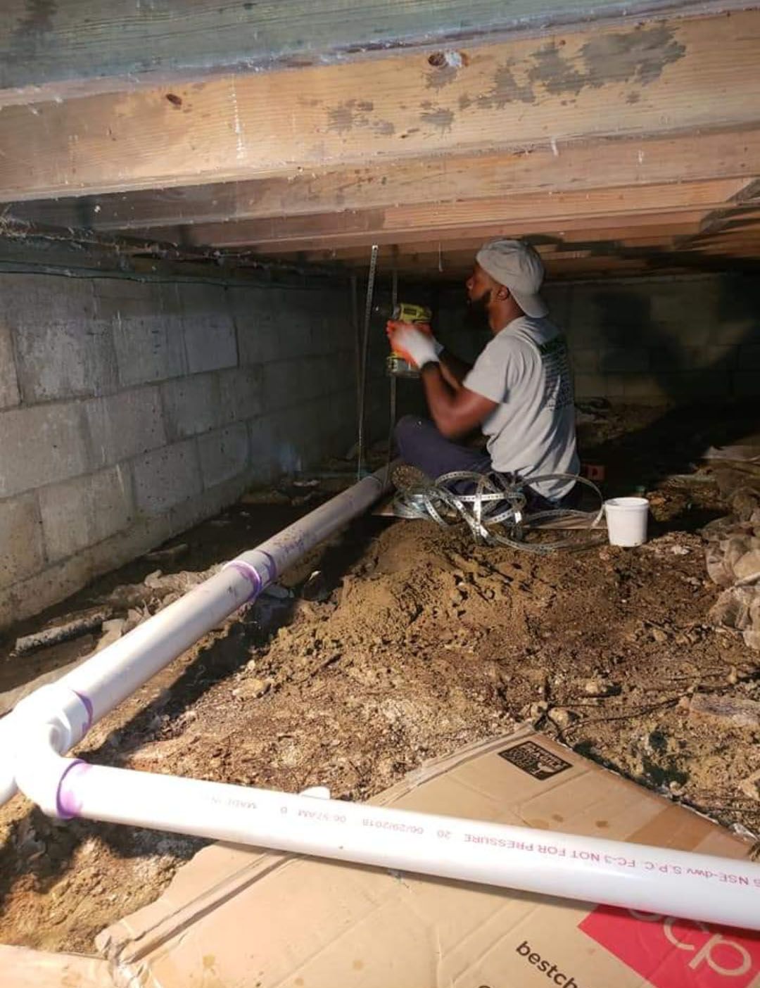 Man seated in a crawl space, working on plumbing with PVC pipes. Soil and cinder block walls surround him.