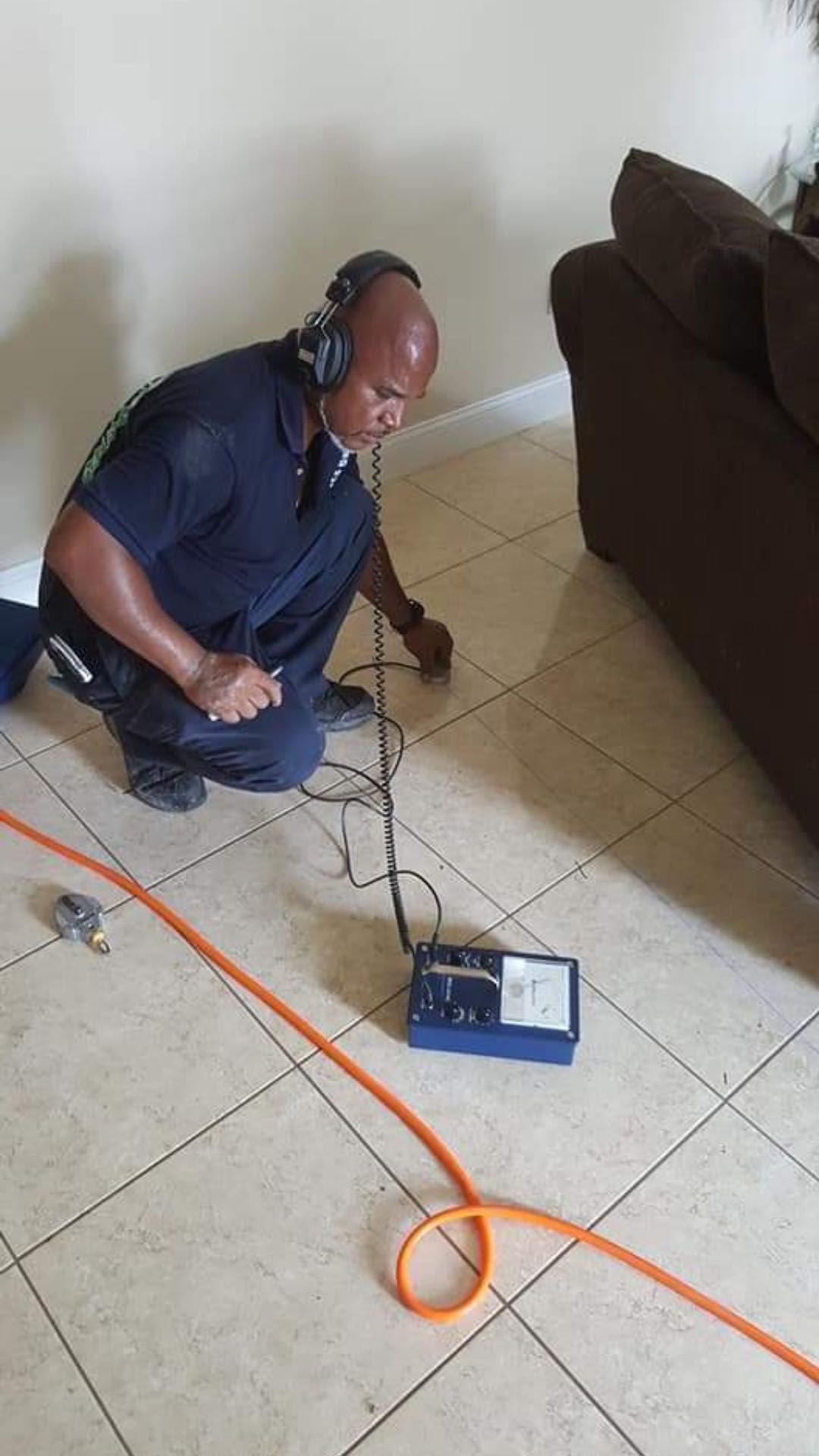 Man using listening device on tiled floor near couch. Orange cable runs to a device.