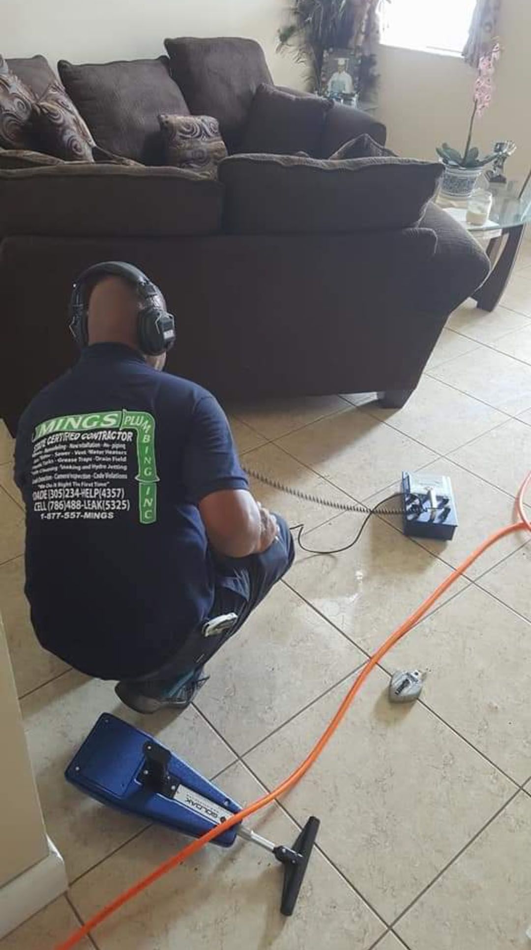 Man with headphones, using equipment on tile floor near a brown couch, likely detecting a leak.